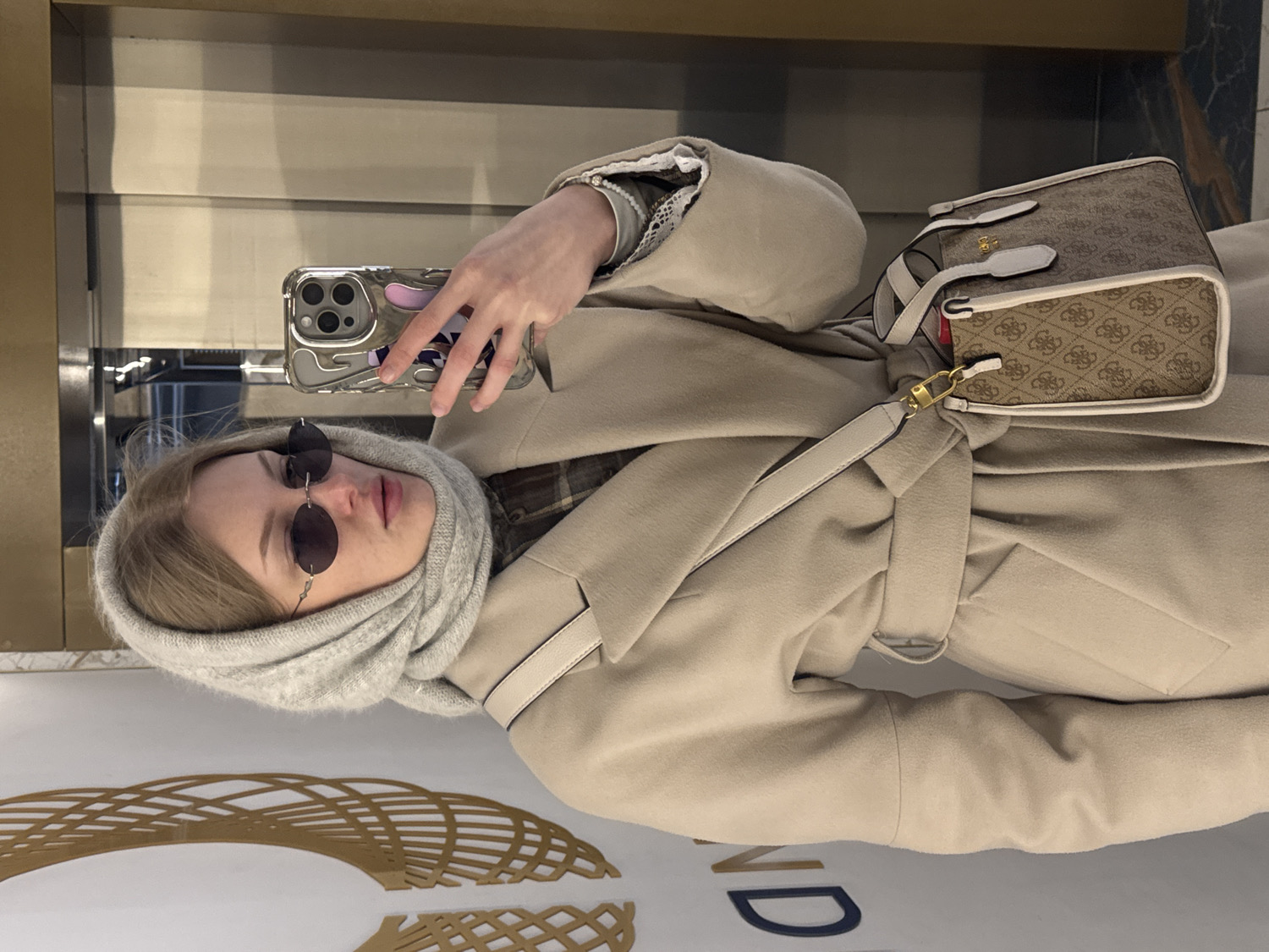 This media file is a mirror selfie featuring a young woman in an indoor setting, likely in Perm, Russia. The image is captured at an angle, with the woman holding a smartphone (appearing to be an iPhone with a clear, textured case) in her right hand, which is reflected in the mirror.
The subject is a young woman with blonde hair, wearing small, round, dark-tinted sunglasses. Her head is covered by a light grey, possibly knitted, hooded scarf or integrated hood. She is dressed in a light beige or tan long coat, cinched at the waist with a tie. A hint of a plaid or checkered garment is visible beneath the coat. On her left wrist, she wears a pearl bracelet.
A small, structured shoulder bag rests against her coat. The bag is light brown and beige with a repeating "Guess" logo pattern, white leather accents, and a gold-toned clasp on its strap, clearly displaying the "GUESS" brand name.
The background, visible through the reflection, shows reflective surfaces and what appears to be a modern interior space with architectural details, including a decorative patterned element in gold and white. Partially visible text in the lower background reads "ND," likely part of a sign.
The overall impression is of a casual yet stylish outfit, suitable for cooler weather, worn by someone taking a self-portrait in a public or semi-public indoor environment.
