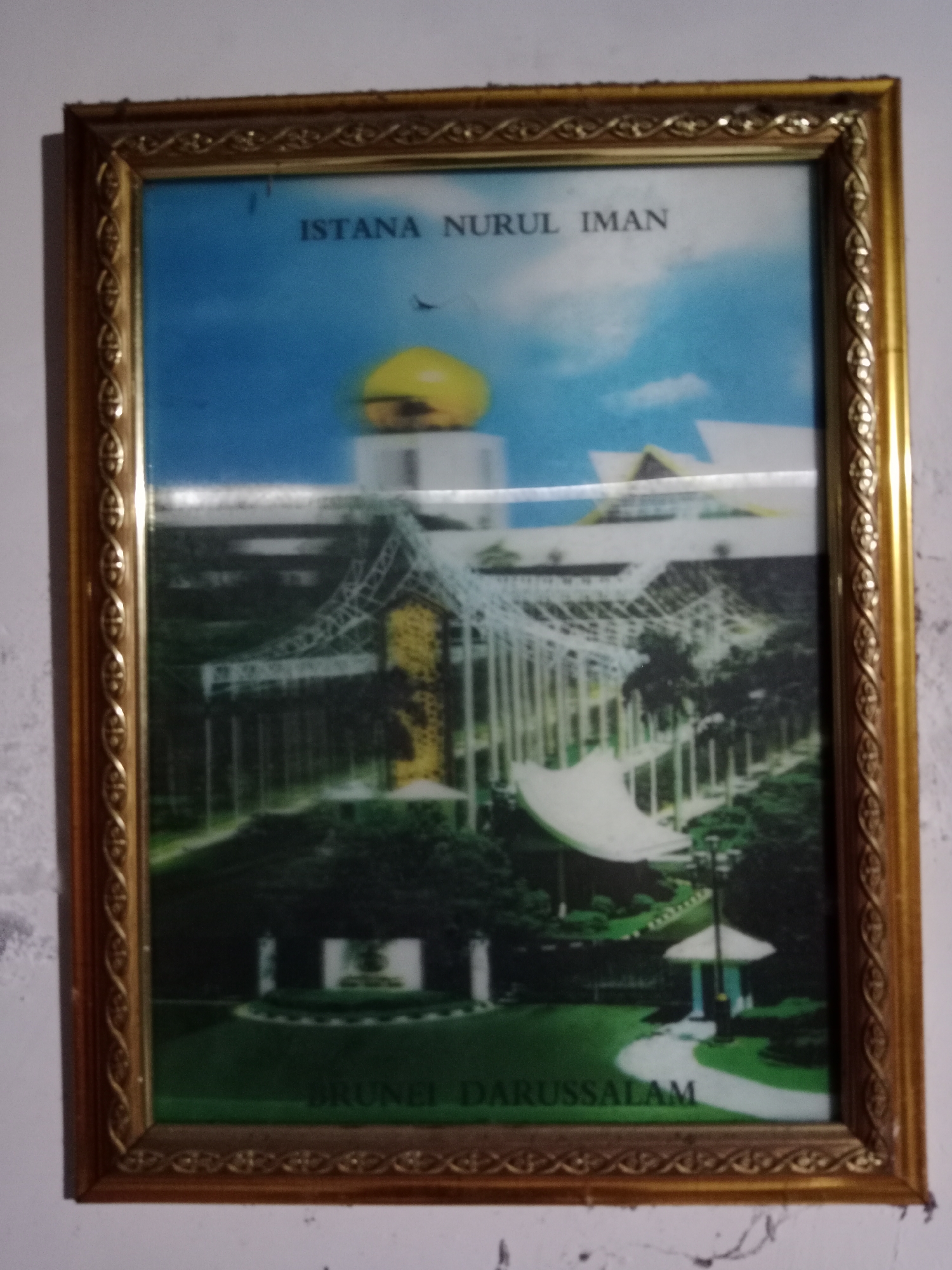 The image is a framed photograph of the Istana Nurul Iman, the official residence of the Sultan of Brunei. The palace is depicted with its distinctive golden dome and grand architecture, set against a blue sky with some clouds. The grounds of the palace are visible, featuring manicured lawns, trees, and pathways. Text at the top of the image reads "ISTANA NURUL IMAN," and at the bottom, "BRUNEI DARUSSALAM." The photograph is housed in a decorative gold frame with intricate patterns along the edges. The image quality is slightly degraded, with a visible grain and some reflections on the surface of the photograph. The photo is hanging on a textured, light-colored wall, likely in a home or office.