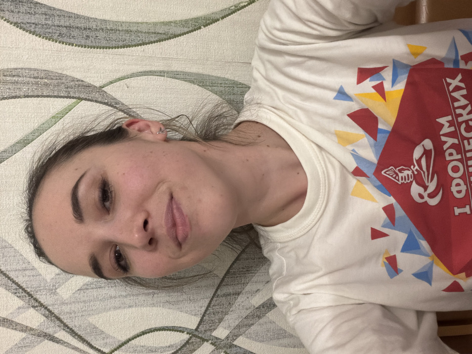 The image portrays a single female subject indoors in Komsomolskiy, Russia. The adult female, with fair skin and dark hair pulled back, occupies the majority of the frame, facing the camera directly with a subtle, closed-mouth smile. She wears a white, long-sleeved t-shirt featuring a prominent graphic design. This design includes a large red trapezoidal area at the bottom, partially obscured, with white Cyrillic text reading "I ФОРУМ ШНЕЖСКИХ" and a stylized white emblem depicting a hand holding papers above a wave-like element. Above the red section, a pattern of blue, red, and yellow geometric triangles is visible. The subject has small silver hoop earrings in both ears. The background consists of a wall covered in patterned wallpaper with interlocking, curvilinear designs in shades of gray and green. A light brown wooden object is partially visible in the lower left corner of the frame.