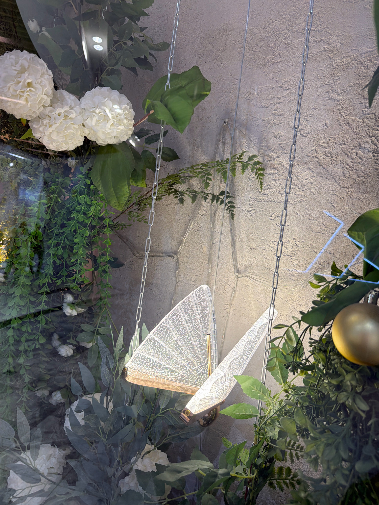 The image shows a decorative display with artificial greenery and white flowers, possibly in a shop window. A large, white, intricate butterfly-shaped light fixture is suspended by chains. Adjacent to the butterfly light is a golden ornament, also appearing to be part of the decoration. The background is a textured, light-colored wall. The overall scene suggests an artistic or festive arrangement. The location is identified as Pavlodar, Kazakhstan.