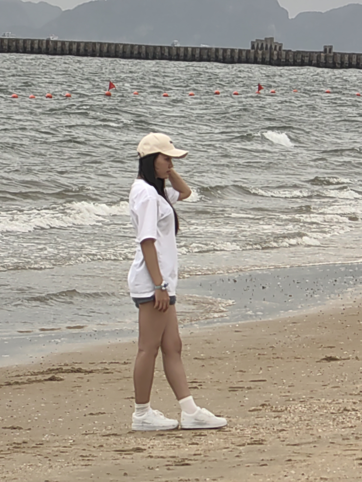 The image depicts a person standing on a beach in Bai Chay Ward, Vietnam. The person is wearing a beige baseball cap, a white t-shirt, denim shorts, white socks, and white sneakers. They are facing to the right, with their left hand raised to their head. The beach is sandy, and waves are visible in the water. A barrier made of red buoys and flags floats on the water. In the background, a pier or seawall extends into the sea, with buildings visible at the end of it. Hills are visible in the far background. The sky is overcast.