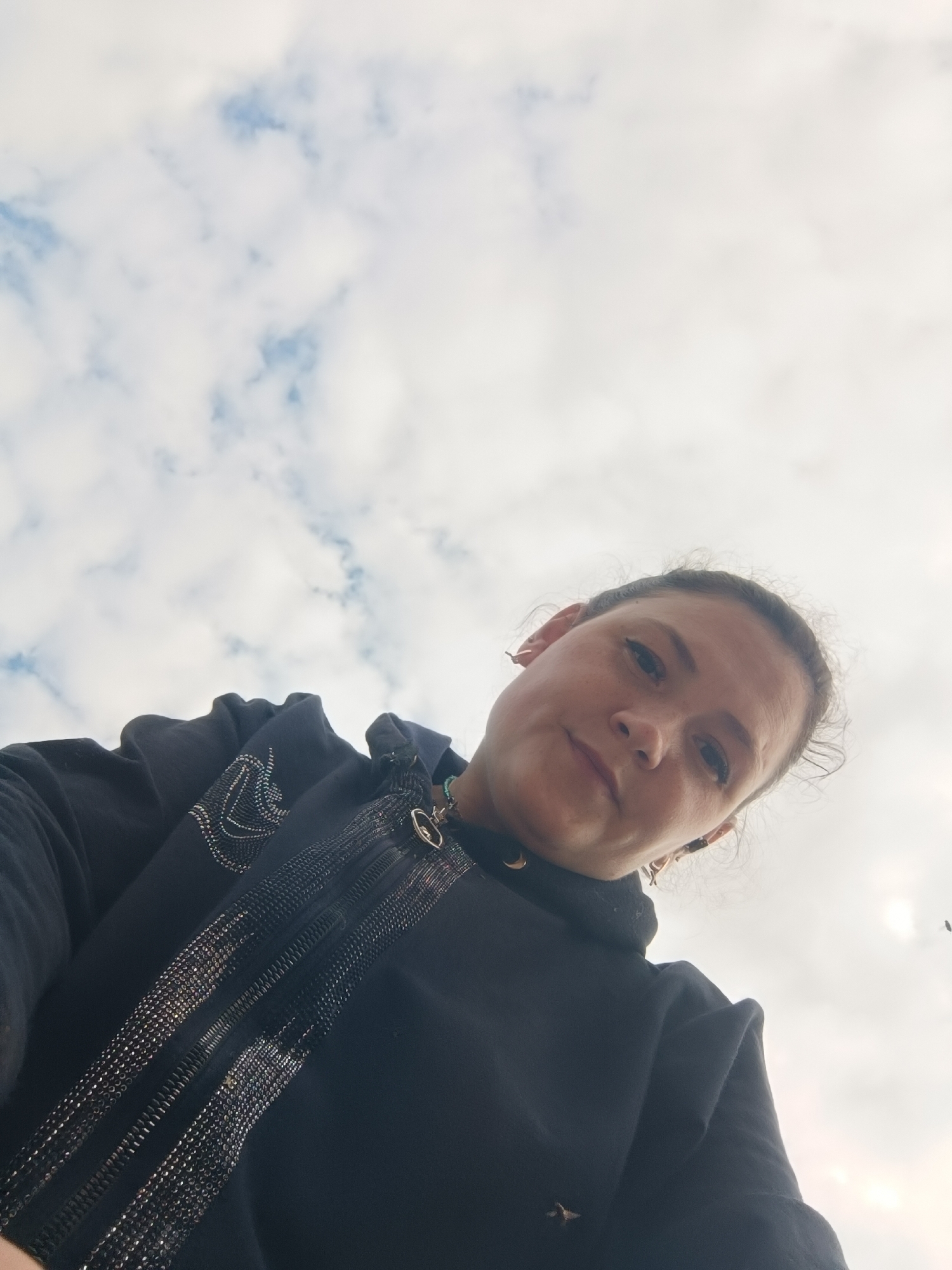 A low-angle, outdoor, waist-up selfie is shown, featuring a woman against a cloudy sky. The woman is wearing a black hooded jacket adorned with reflective detailing along the zipper and on the upper chest. She has dark hair pulled back and is looking downwards at the camera with a neutral expression. Two small birds are visible in the distance against the sky. There is no indication of the city of Karsha, Russia in this image.