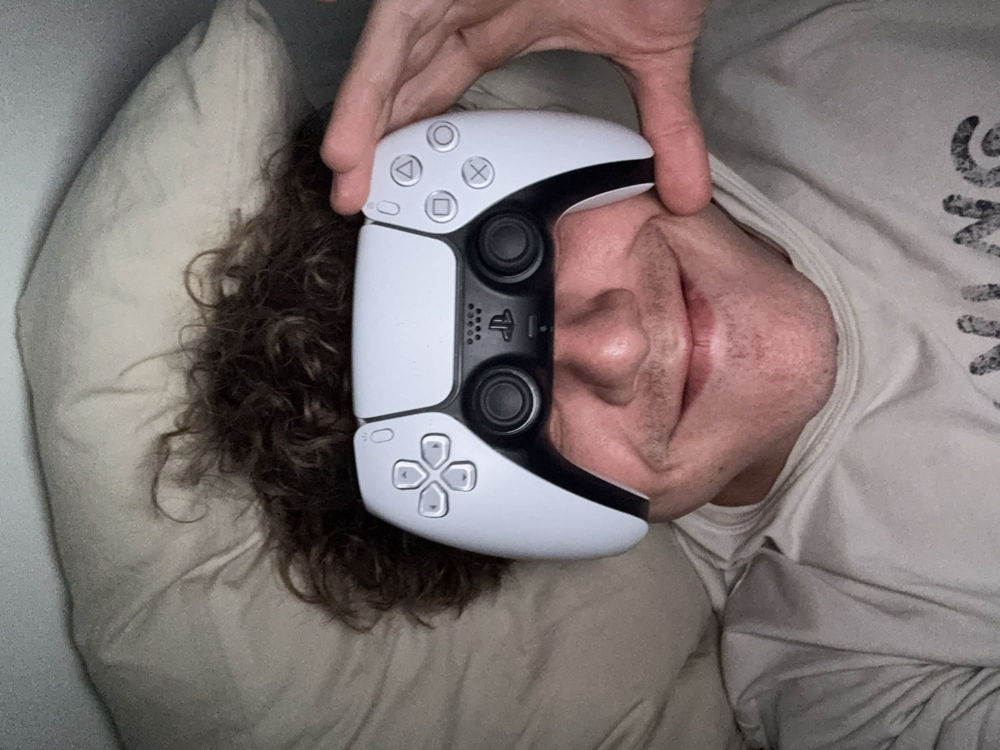The image shows a person lying on a pillow, with a PlayStation 5 DualSense controller held over their face. The person has curly hair and is wearing a light-colored t-shirt. The background is a light-colored wall and pillow. The location is not discernible from the image.