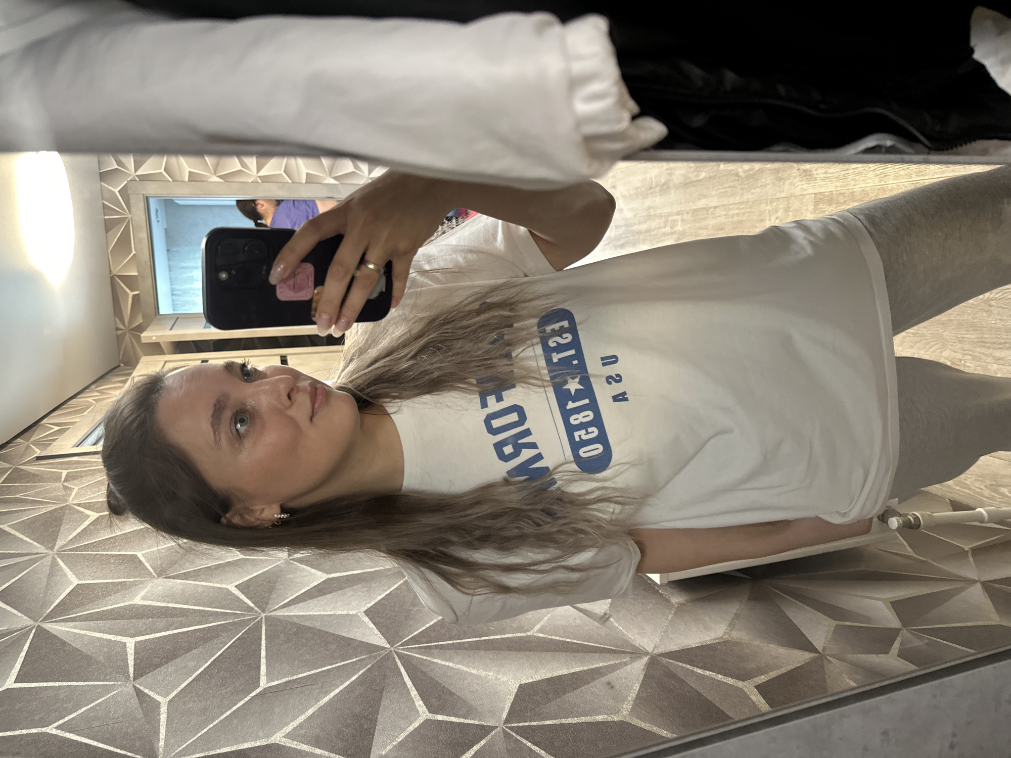 A young woman is taking a mirror selfie indoors. She is wearing a white t-shirt with blue lettering that reads "FORM EST. 1850 USA" and light grey sweatpants. She has blonde hair tied up in a bun and is wearing hoop earrings and a ring on her right hand. The background features a geometrically patterned wallpaper and a wooden floor. In the reflection, a door is visible, leading to another room where a person wearing a purple shirt can be seen. The setting appears to be a domestic environment, possibly a hallway or a room with a large mirror. The location is identified as Voznesenka, Russia.