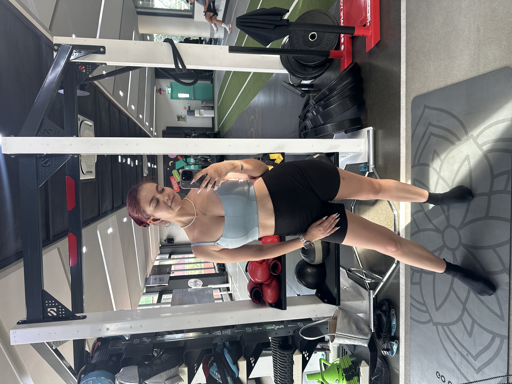 A young woman with red hair pulled back stands in a gym, taking a selfie in a mirror. She wears a light blue sports bra, black athletic shorts, and black socks. Around her neck is a pearl necklace. In the background, gym equipment is visible, including weight racks, boxing gloves, and weight plates. The gym appears to be a modern facility with athletic turf flooring and bright overhead lighting. The environment suggests an indoor fitness space.