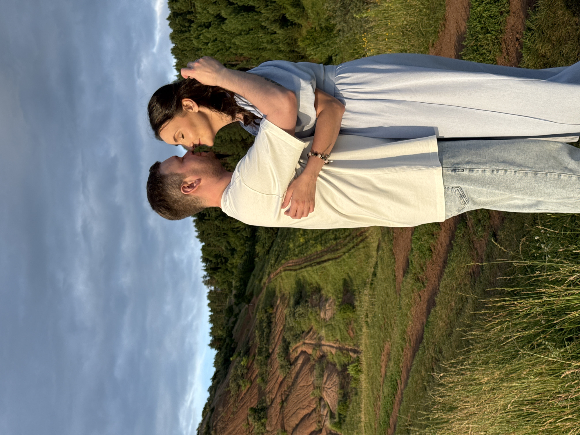 A man and a woman are embracing outdoors, their foreheads touching and eyes closed. The man, wearing a white t-shirt and light blue jeans, has his arms around the woman. The woman, dressed in a light blue dress, has her left arm around his neck and her right hand in his hair. Both have their eyes closed and appear to be sharing an intimate moment. The background features a hilly landscape with patches of green grass and trees. A dirt path winds through the grassy hills. The sky above is cloudy. The location is likely a rural or natural area, possibly on the outskirts of Olkhovka, Russia, given the terrain and vegetation.