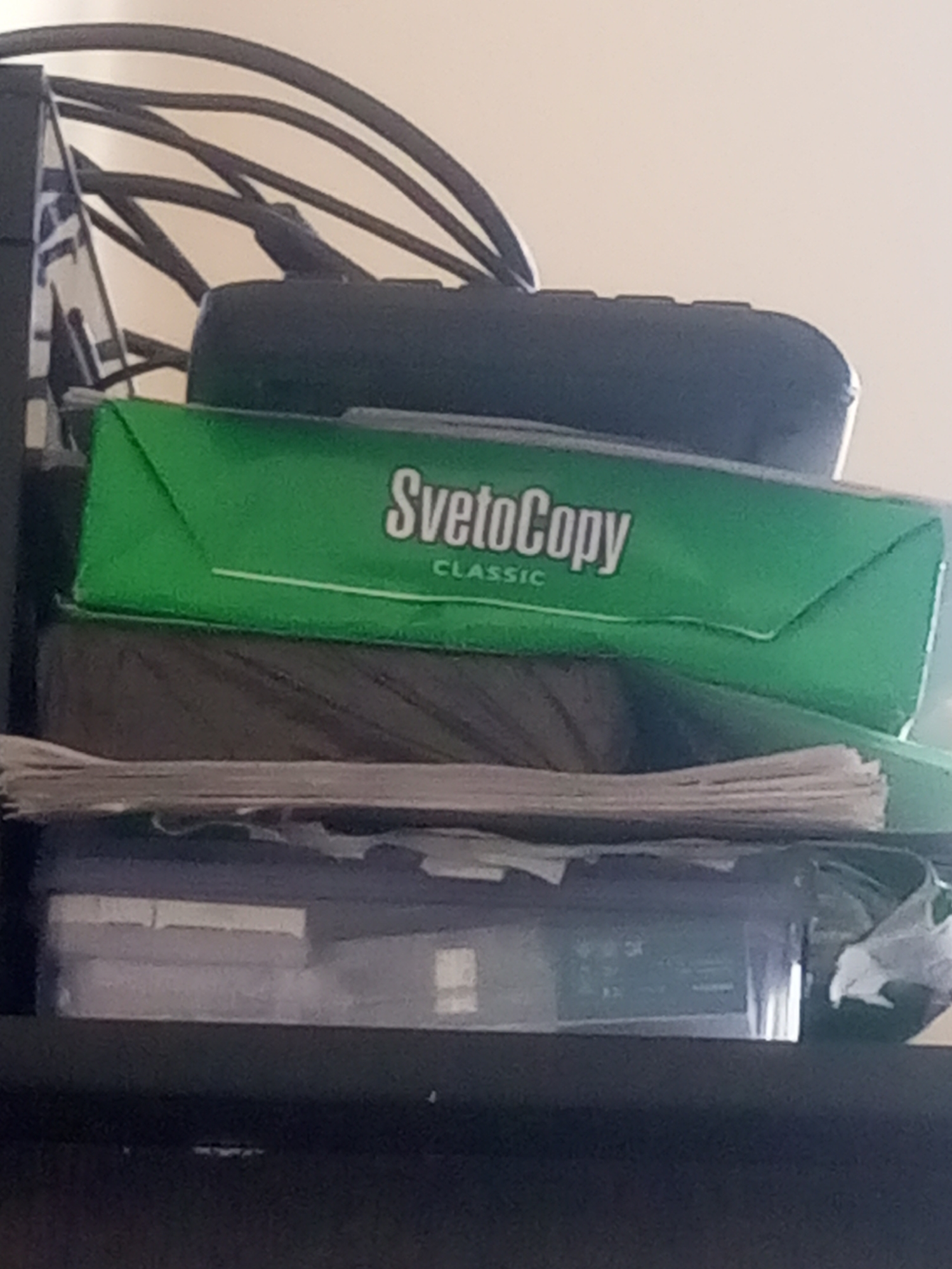 This is a close-up, vertically oriented, medium-shot photograph of a stack of items on a shelf. The dominant object in the foreground is a green ream of paper with "SvetoCopy CLASSIC" printed in white on it. This paper ream is placed on top of a dark, fibrous material, possibly a bundle of yarn or thread, which in turn rests on a stack of what appears to be loose papers. Below this are two stacked translucent plastic containers, the top one filled with papers and the bottom one appearing empty. In the background, a black, curved object, possibly part of a computer monitor or a stand, is visible with several black cables running through it. The lighting suggests an indoor environment, likely a room, with a plain, light-colored wall in the background. The image does not contain any specific indicators of the city of Bokhtar, Tajikistan.