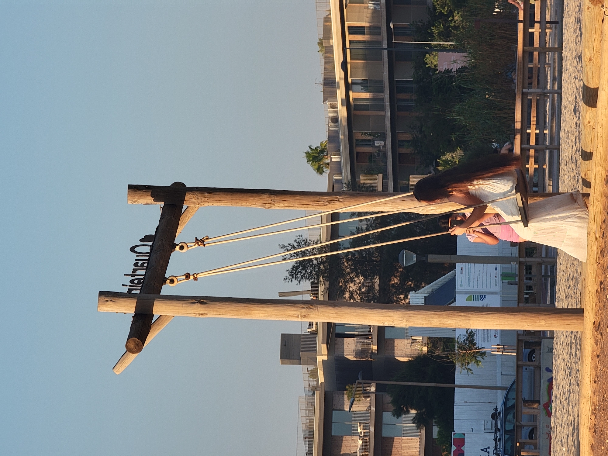 A side profile, medium shot captures a young woman with very long dark hair seated on a wooden swing. She is wearing a white strapless dress. To her left, partially obscured by the swing structure, another individual, a young girl, is visible wearing a pink shirt and holding a phone up. The swing set is constructed from rough, natural wood posts and beams, with the ropes of the swing attached to a horizontal beam. A metal sign with black lettering, appearing to read "QUARTEIRA," is affixed to the top beam. The background consists of modern, multi-story buildings with balconies and glass windows, characteristic of a coastal urban environment. Trees and dense vegetation are interspersed between the buildings and the foreground. The ground in the foreground is sandy, suggesting a beach or park setting. The scene is illuminated by the warm, low-angled light of what appears to be late afternoon or early evening.