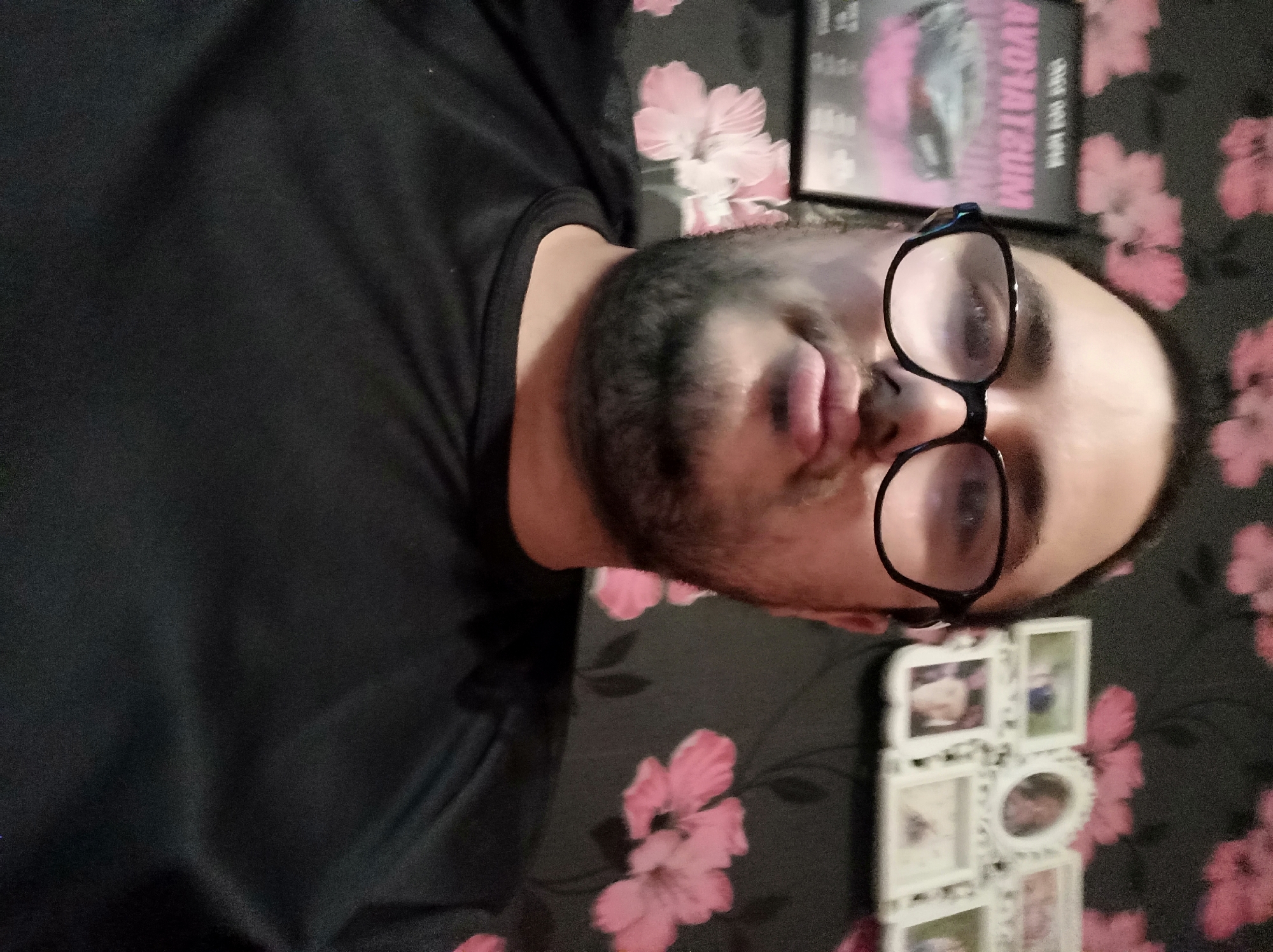 A close-up, slightly tilted image shows a person wearing glasses with a black frame. The person has a beard and is wearing a dark-colored shirt. The background consists of a black surface with pink flower patterns. A white photo frame with multiple small pictures is visible towards the bottom right. A movie case is seen at the top of the image. The location is implied to be the city of Unknown, Unknown.