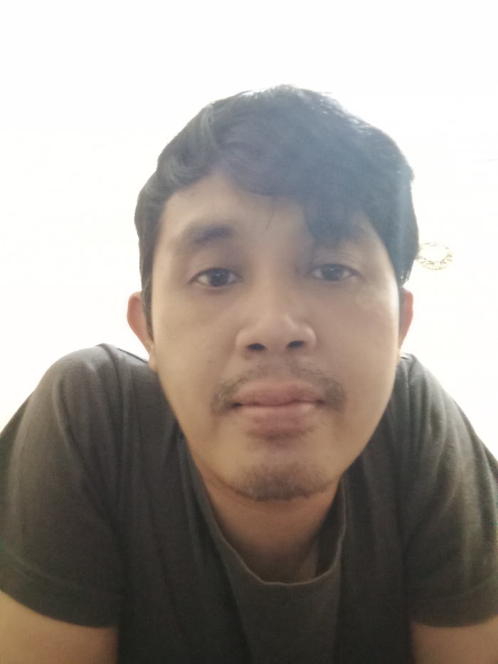 A close-up shot of a man with dark hair and a mustache. He is wearing a gray shirt. The background is a plain white wall. His gaze is directed towards the camera. The lighting is somewhat soft and even, though there's a bright spot above his head. He appears to be positioned slightly below the camera. The overall impression is an informal, unfiltered self-portrait.