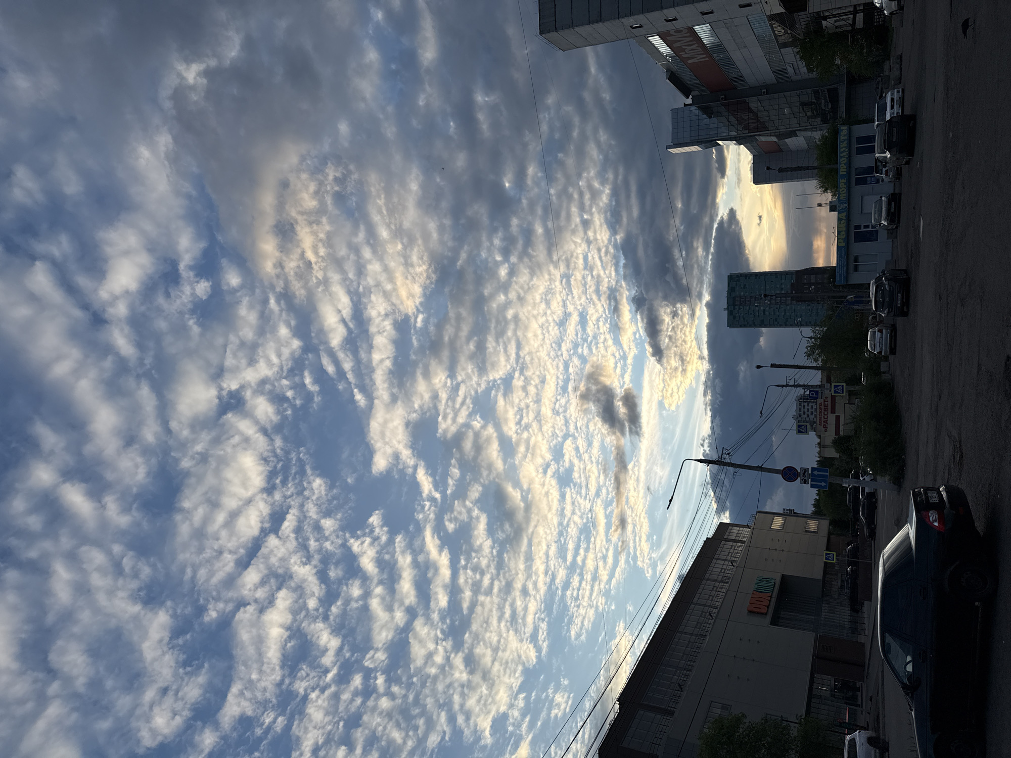 The image shows a street scene in Krasnoyarsk, Russia. The sky is filled with altocumulus clouds, some backlit by the setting sun. Along the right side of the frame are buildings, parked cars, and street infrastructure (poles, wires, traffic signs). In the lower right corner, part of a dark-colored car is visible. Buildings appear to be a mix of commercial and residential, with signs in Russian.