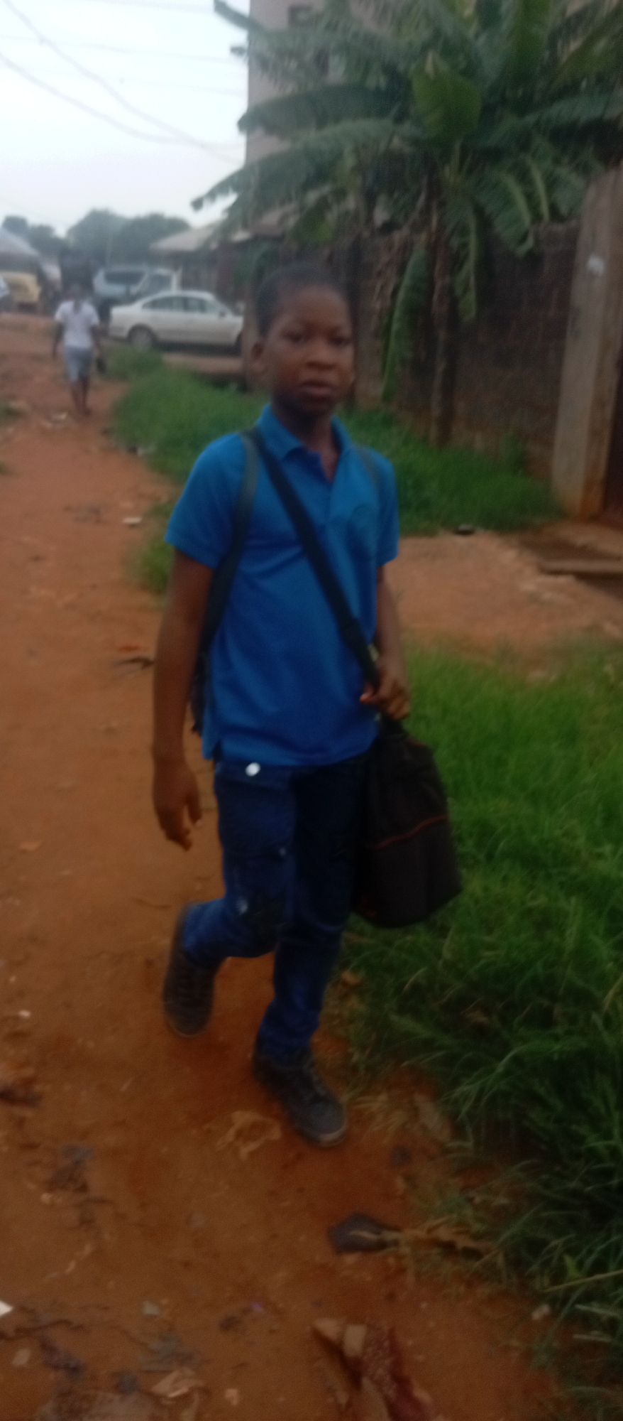 The image shows a young person walking on a dirt path. The person is wearing a blue shirt, dark jeans, and dark shoes, and carrying a black bag. They are walking towards the viewer. In the background, there is a second person and cars. A building and tropical vegetation are visible, including what appears to be a banana plant. The dirt path is bordered by green vegetation. The location is likely in a residential area of Ogidi, Nigeria.