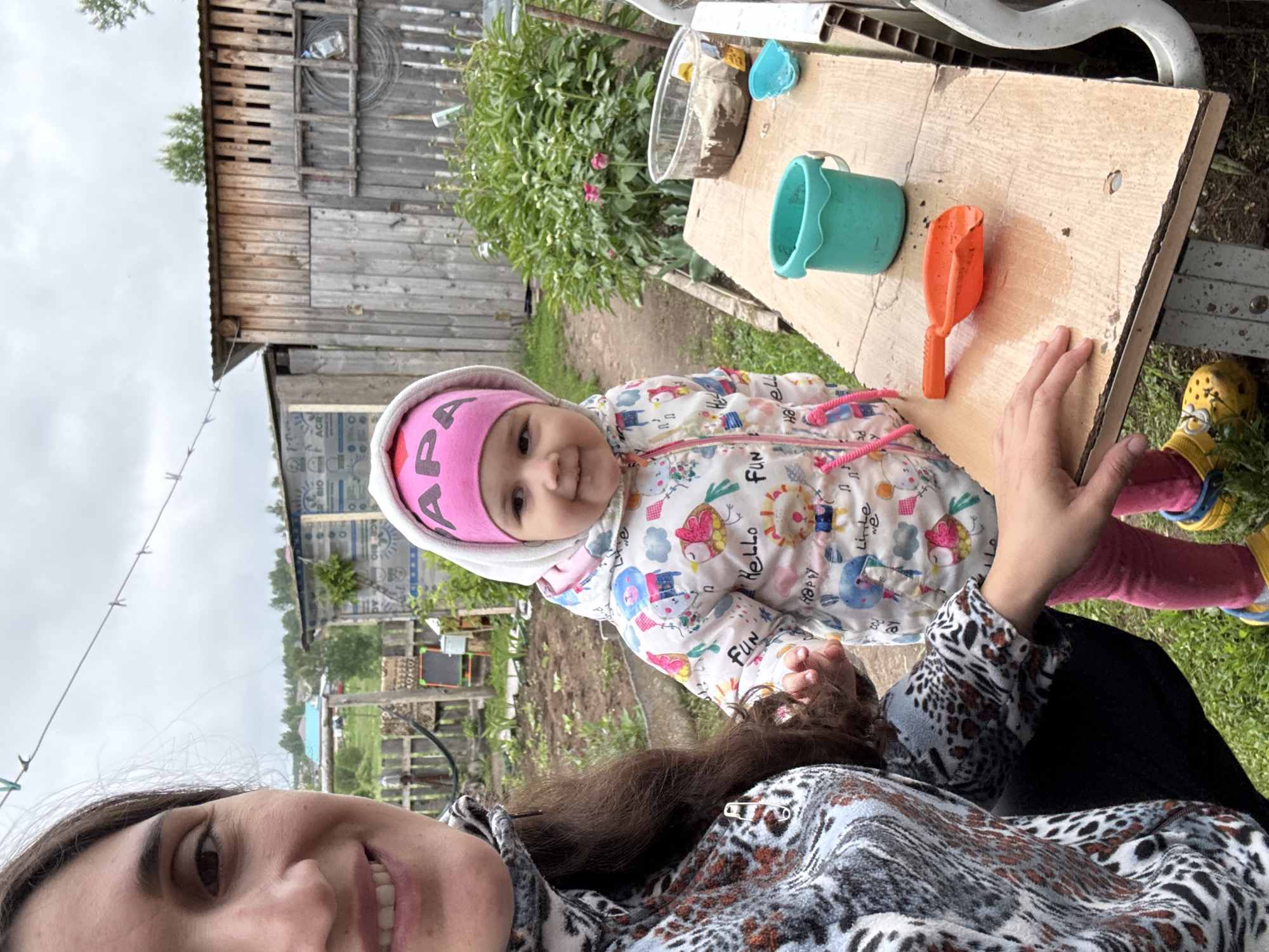 This image captures a candid outdoor moment in Уральское, Russia, featuring an adult taking a selfie with a young child. The perspective is from the adult looking down towards the child and the ground.
The primary subjects are a toddler and a partially visible adult. The child, situated centrally, looks directly at the camera with a pleasant, curious expression. They are dressed warmly in a white hooded jacket adorned with colorful cartoon animals (birds, lions, clouds) and English words such as "HELLO," "FUN," and "HAPPY." A pink hat covers their head, clearly displaying the Cyrillic text "ПАПА" (PAPA), meaning "Dad." They wear pink pants and yellow Crocs-style shoes with blue accents.
The adult, visible in the lower left, has long dark hair and a smiling face. They are wearing a patterned fleece-like top in shades of grey, black, and brown, with a visible safety pin on the chest. One of the adult's hands is near a rustic wooden table positioned to the right of the child.
On the wooden table are several items for child's play: a clear plastic container filled with dirt or mud, a small teal plastic bucket, and an orange plastic spade.
The setting appears to be a rural or semi-rural garden or yard. In the background, there is a large, weathered wooden shed or building with horizontal planks, suggesting an older structure. Lush green foliage, including bushes with some pink flowers, lines the background. The ground is visible as dirt or earth. A clothesline with pegs stretches across the upper left of the frame. The sky is overcast and uniformly gray, indicating a cloudy daytime.
The overall impression is one of a cheerful and casual outdoor activity, possibly involving mud play, with a clear sense of affection between the adult and child.