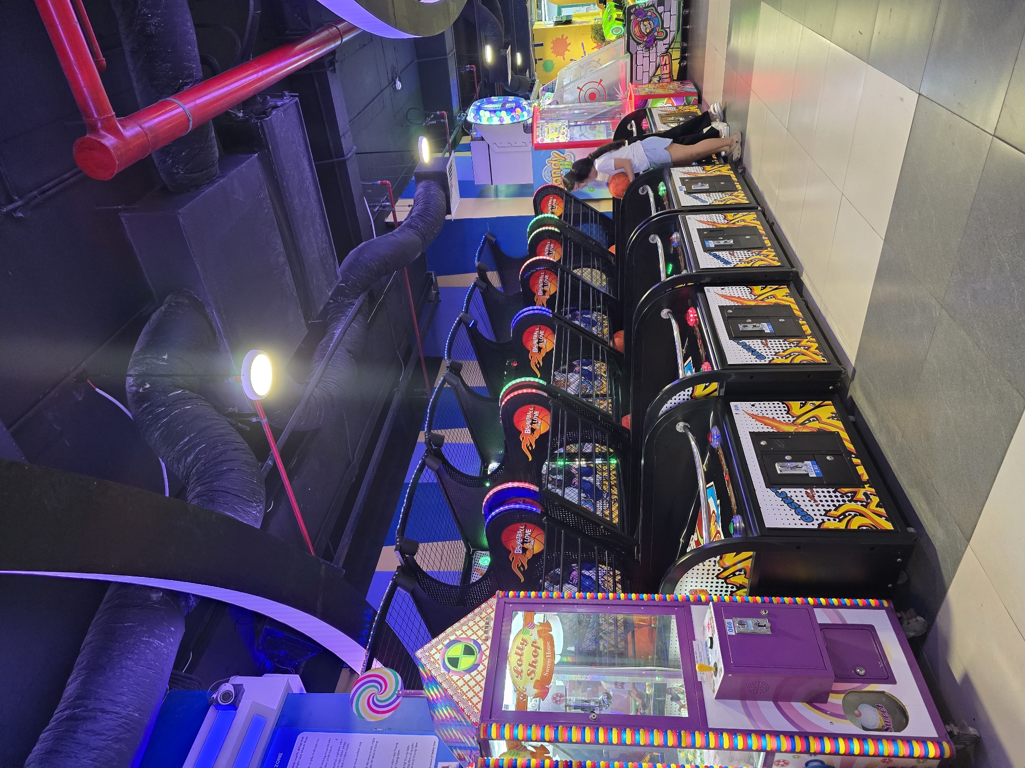 This image depicts an indoor arcade in Nha Trang, Vietnam. The scene is dominated by a row of basketball arcade games, each with colorful lights and graphics, some displaying "Basketball Love." A girl in a white t-shirt and blue shorts is playing one of the games, holding a basketball. To the left of the arcade games is a purple and rainbow-striped claw machine labeled "Lolly Shop Sweety House." In the background, there are more arcade machines, including one with a target and a cartoon character. The environment features a dark ceiling with exposed pipes and ventilation ducts, and brightly lit signs and game displays. The floor is tiled in shades of gray and white. The overall atmosphere is that of a lively entertainment venue.