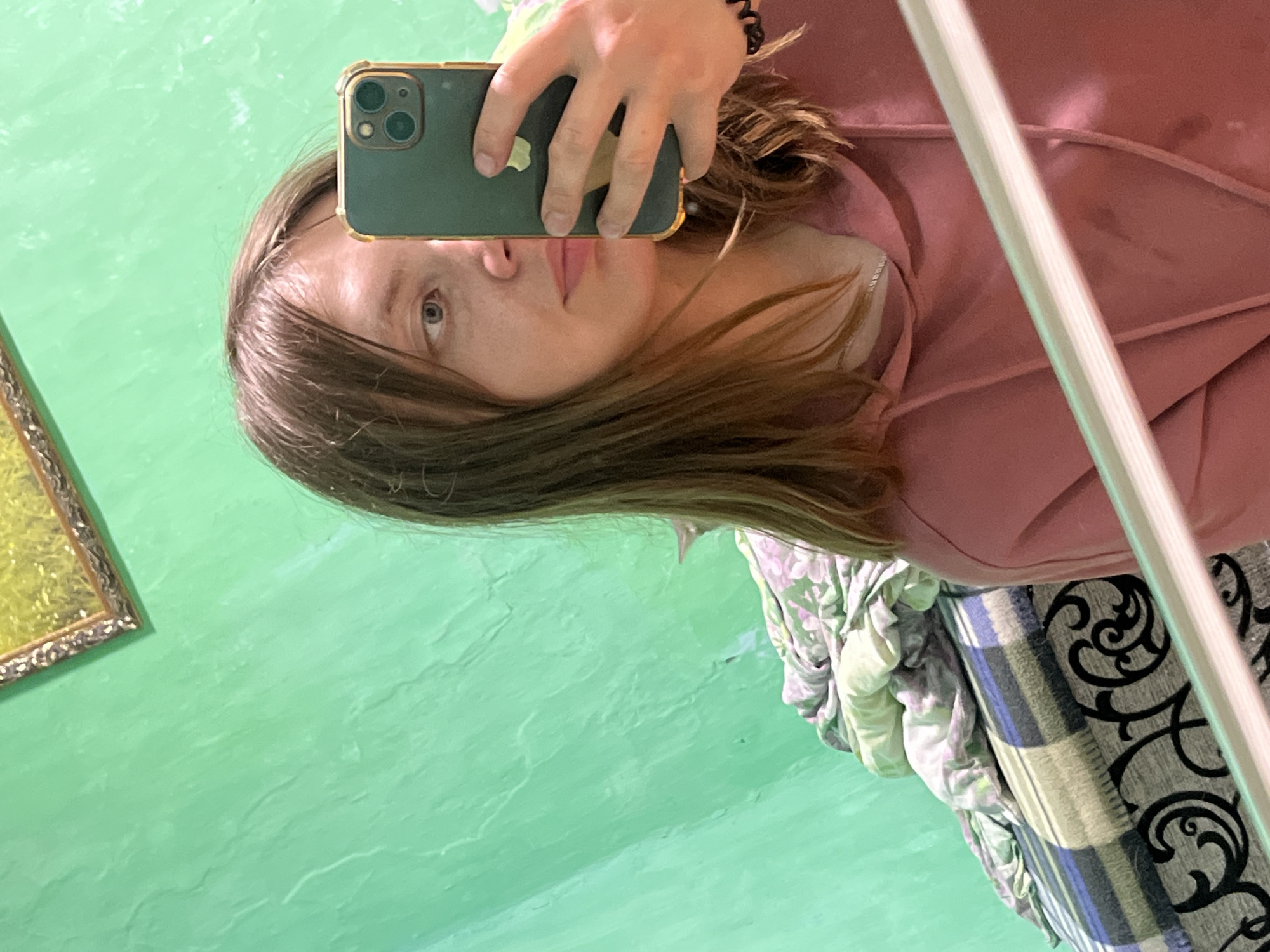 The image is a selfie taken by a young woman with shoulder-length brown hair. She is wearing a pink hooded sweatshirt and is holding a dark green smartphone in her right hand to take the picture. Her left eye is visible, and she is looking at the camera. The background is a textured green wall, with a framed picture partially visible in the upper left corner. To the left of the woman, a bed is visible, covered with a patterned duvet and a striped blanket. There is no identifiable information in the image to suggest the location in the city of Nizhny Tagil, Russia.