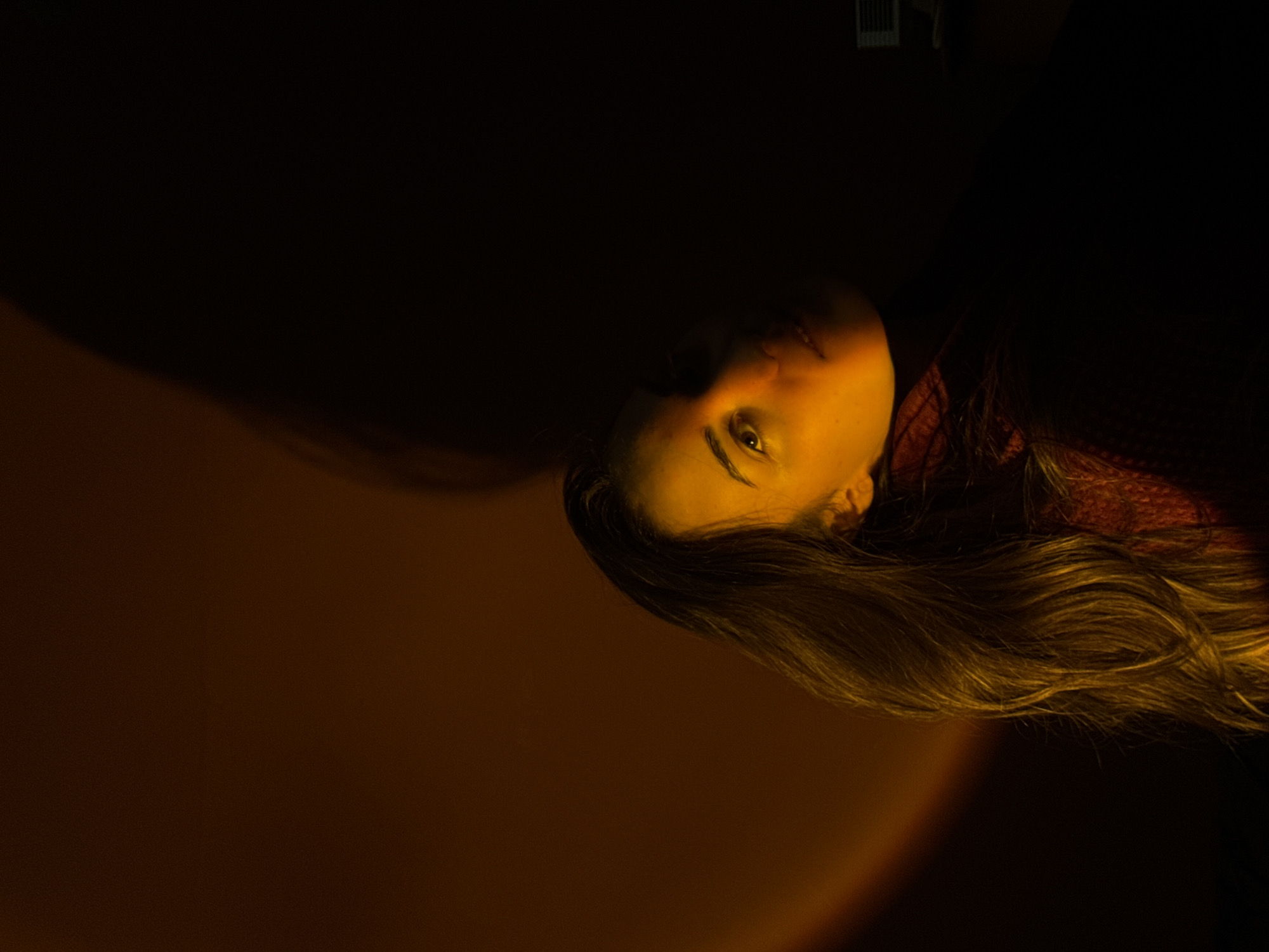 A dimly lit, rotated close-up photo shows a person with fair skin and long, dark blonde hair. The person is wearing a dark-colored top. The lighting casts a yellow hue on the person's face, and their left eye is visible. Part of a red garment is visible near the person's shoulder. A shadow stretches across the left side of the image. An object resembling a vent is faintly visible in the top right corner. The location, Moscow, Russia, is not evident in the image.
