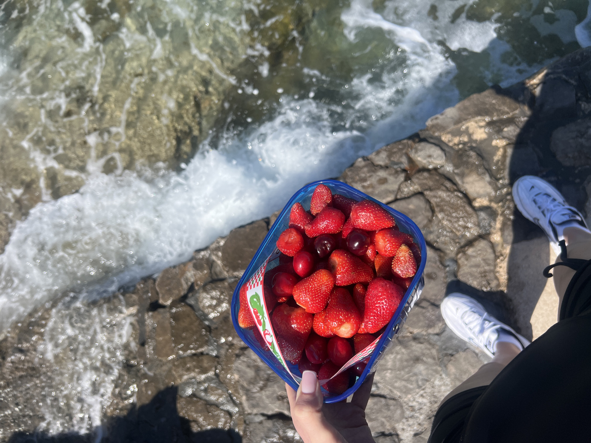 A person is holding a container of strawberries and cherries on a rocky outcropping in Prijevor, Montenegro. The sea is visible in the background. The person is wearing white shoes and black shorts.