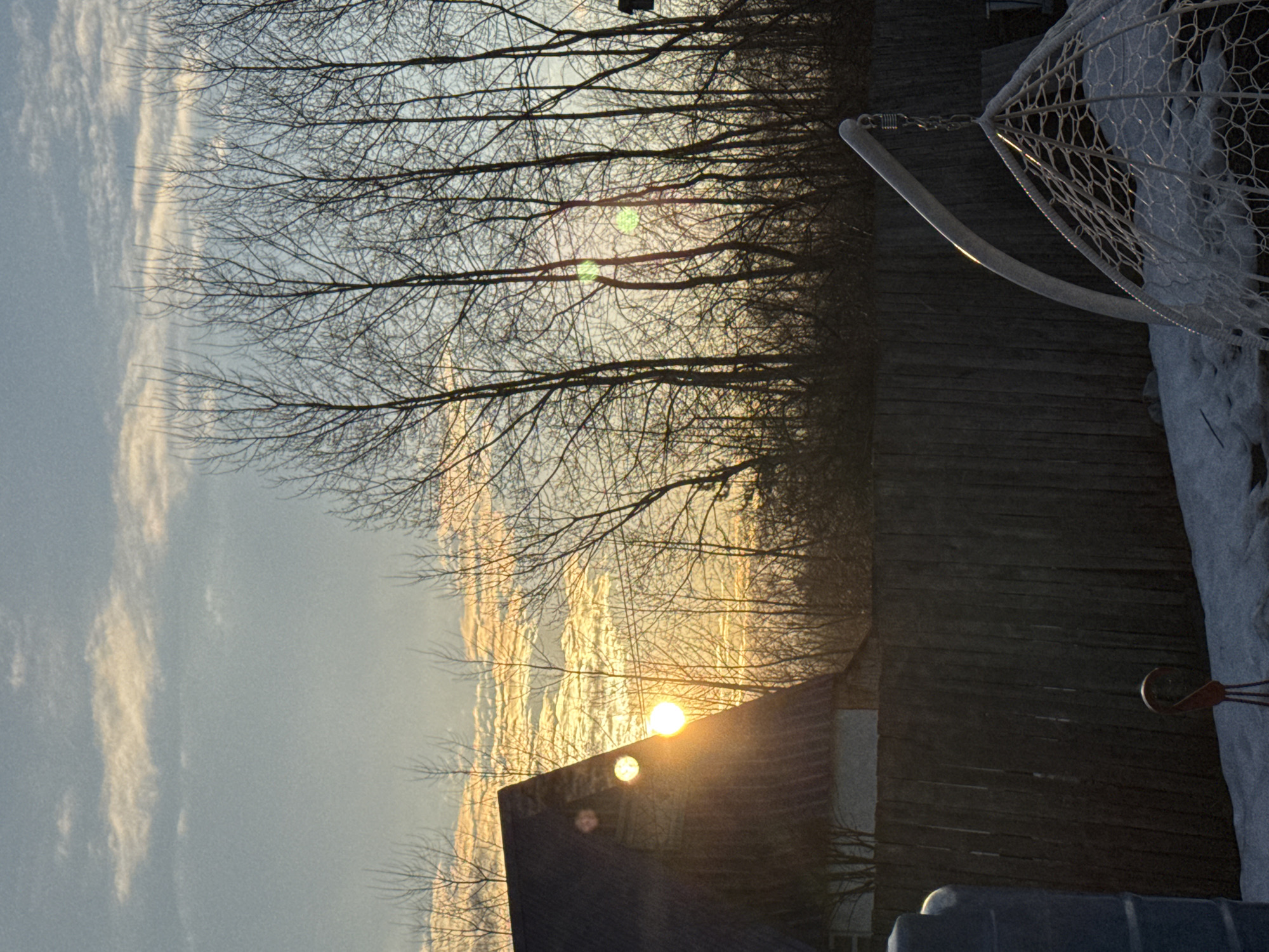 The image shows a winter scene. Bare trees are silhouetted against a bright, cloudy sky with a sunset. The sun is partially obscured by a building roof, causing a lens flare. A wooden fence and a snow-covered basketball hoop are visible on the right. A dark-colored object is partially visible at the bottom of the frame, partially obscured by shadows. The location is Chaikovsky, Russia.