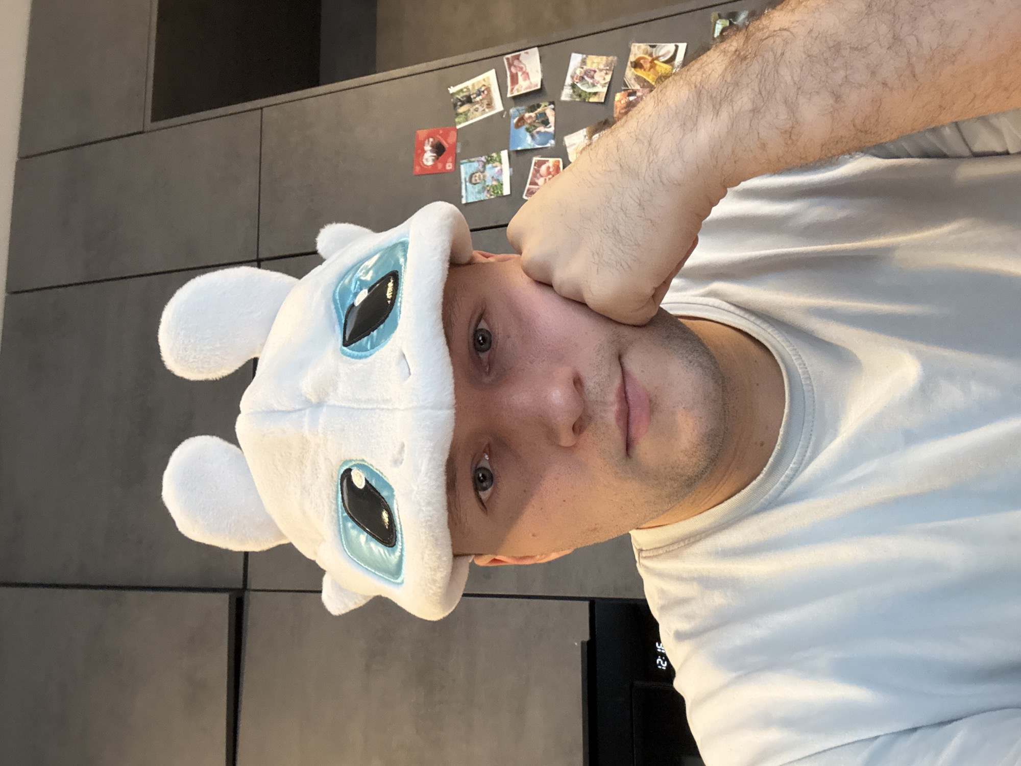 A man is pictured from a close, angled perspective, wearing a white plush hat with blue-and-black cartoon eyes and small ears on top. His right fist is positioned against his head. He is wearing a light-colored T-shirt. Behind him is a gray wall with a grid-like pattern, and several photographs are affixed to the wall. A black appliance is also visible in the background. Based on the evidence available, there is insufficient information to conclude the photograph was taken in the city of Krasnoyarsk, Russia.