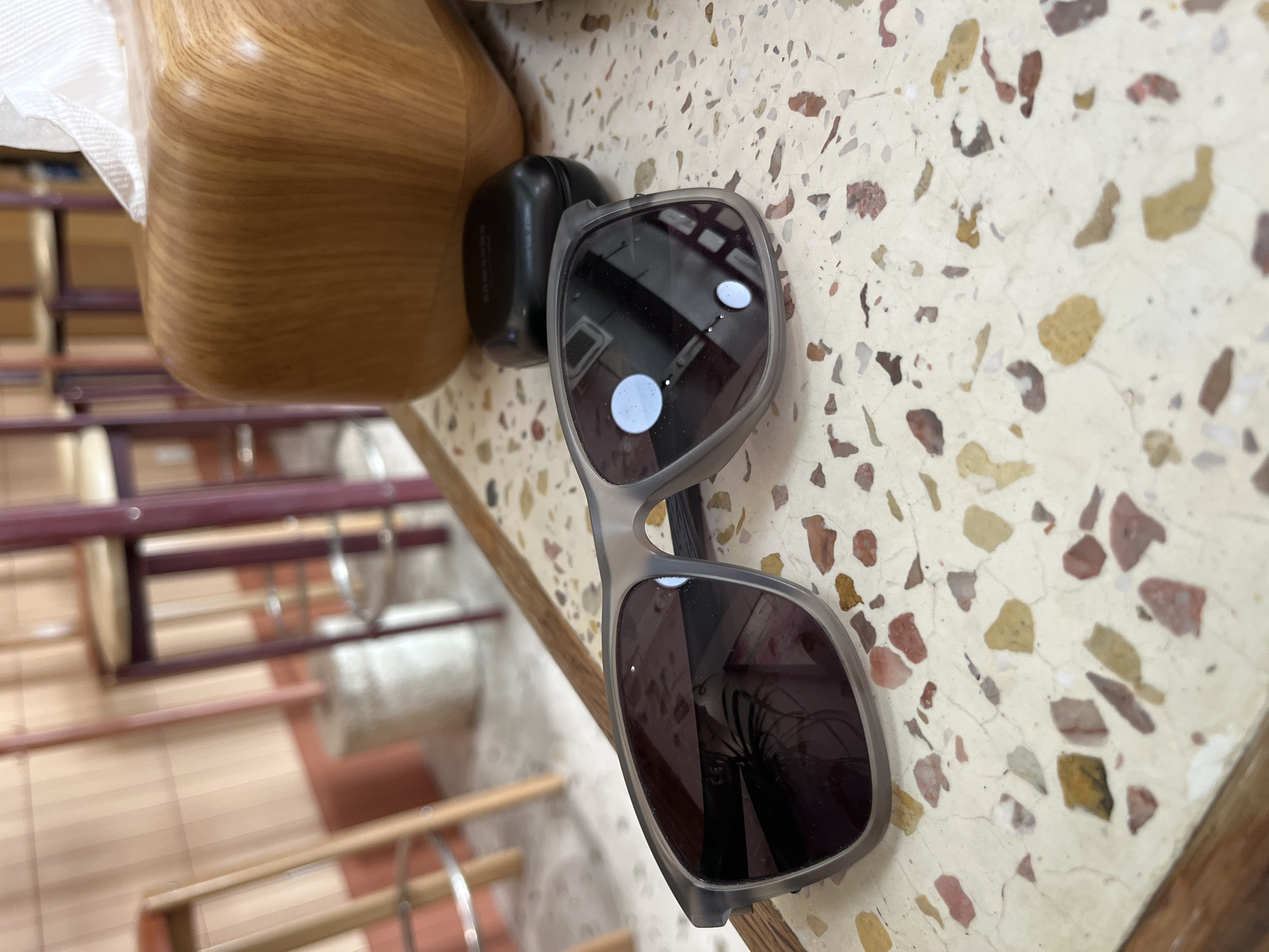 The image shows a close-up view of a table in what appears to be an indoor space, possibly a café or restaurant in the city of Bac Nha Trang Ward, Vietnam. A pair of sunglasses with a dark gray frame lies on a light-colored terrazzo tabletop. Next to the sunglasses are a dark-colored earpiece case and a wooden tissue holder with white tissue. In the blurred background, there are wooden chairs and what appears to be a light-colored floor. The angle of the shot is slightly tilted.