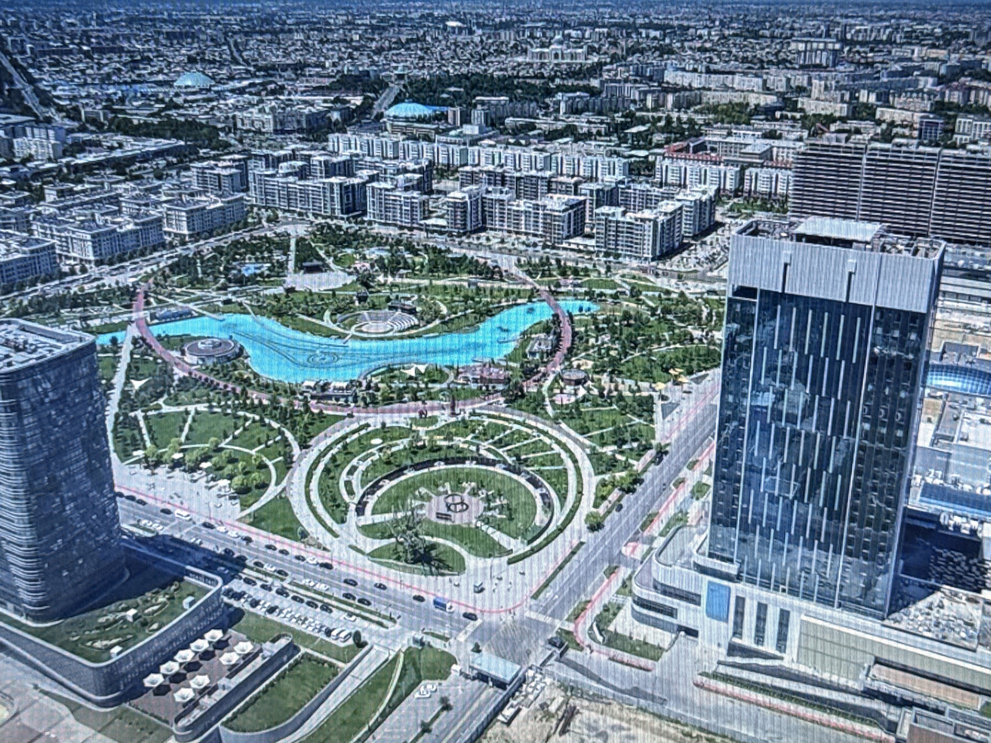 This aerial view depicts a modern cityscape characterized by a large, man-made lake or river, surrounded by lush green parkland. Two prominent skyscrapers, one on the left and another on the right, anchor the scene, their glass facades reflecting the daylight. The park features winding paths, circular garden layouts, and what appears to be an outdoor amphitheater or stage. Beyond the park, a dense urban sprawl of residential and commercial buildings stretches into the distance, interspersed with trees. Roads with light traffic, including cars and buses, are visible throughout the urban area and around the park. The overall impression is one of a well-developed urban environment with significant recreational green space. The time of day appears to be daytime, with clear weather, as indicated by the lighting and absence of clouds. The image provides a broad overview of the urban planning and infrastructure of the location. There is no visible text in the image.