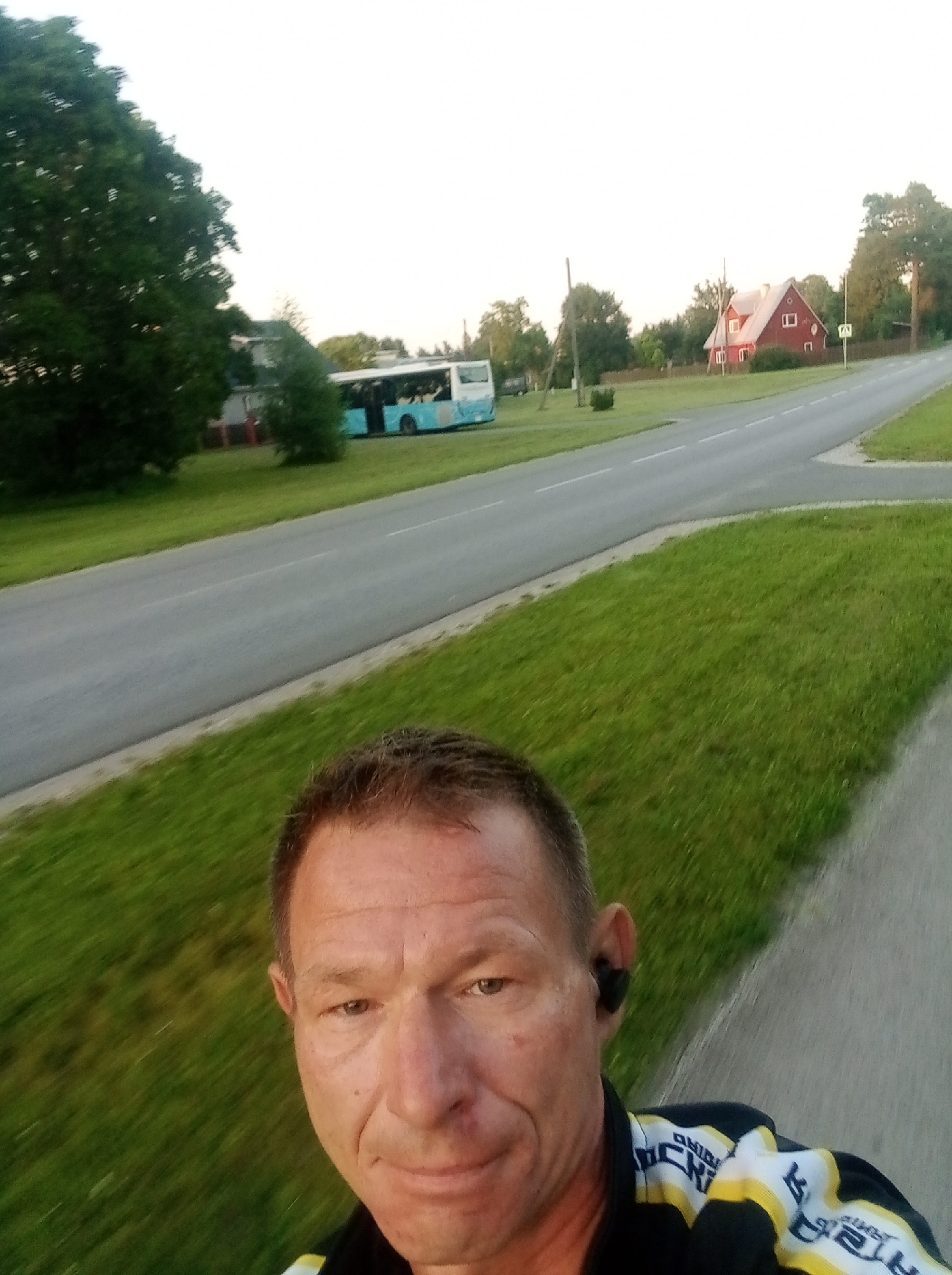 The image shows a selfie taken by a man outdoors, likely in the city of Tapa, Estonia, during daylight hours. The man is positioned in the foreground, with his face partially visible and turned towards the camera. He is wearing a black jacket with yellow and white accents and black earbuds. Behind him, a paved road runs diagonally across the frame, bordered by a grassy verge. Further in the background, a blue and white bus is visible on the side of the road, parked near a building. To the right of the bus, a red-roofed house stands with trees surrounding it. The sky is overcast. The man appears to be on a sidewalk or path adjacent to the road.