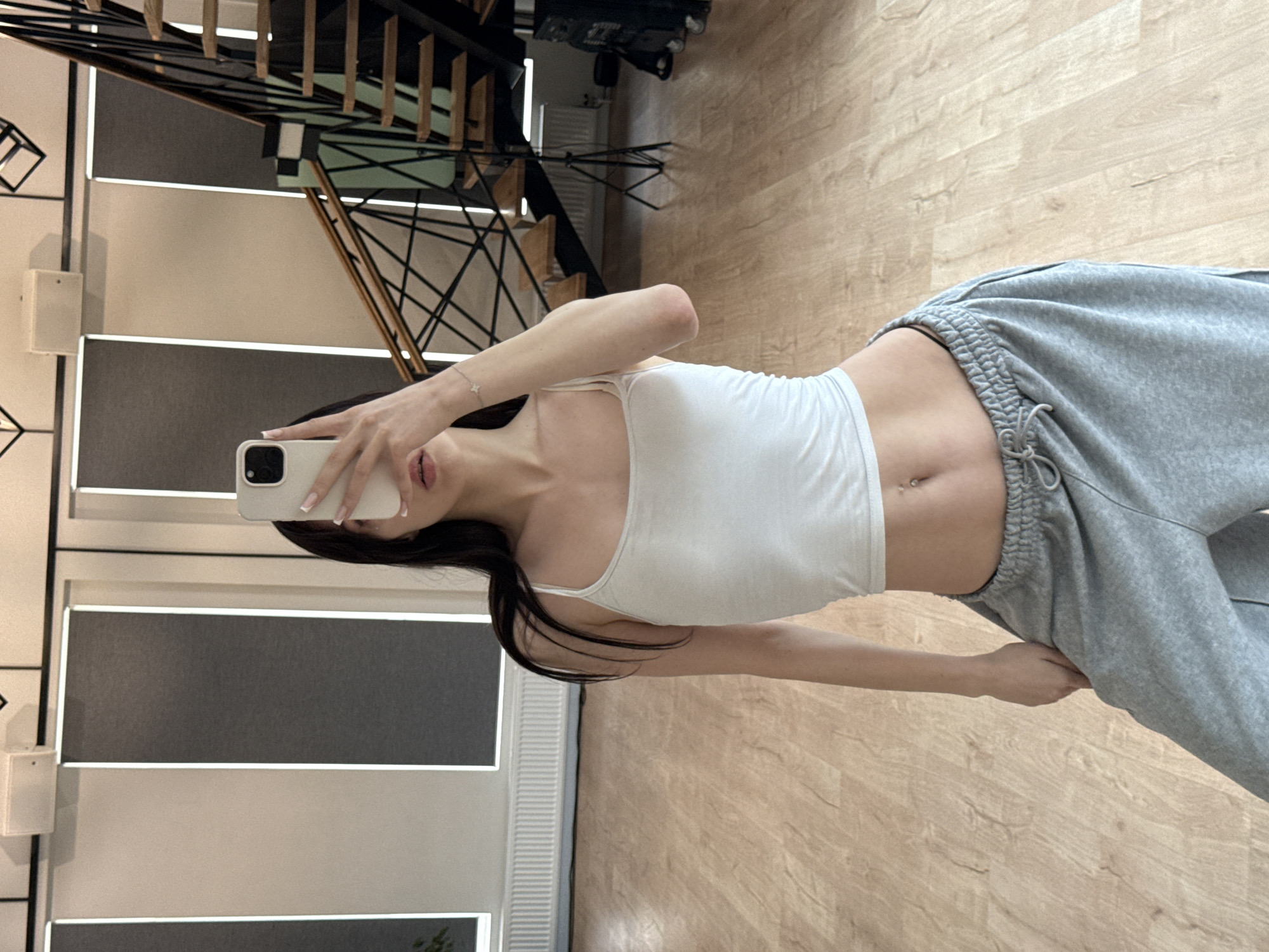 A young woman, with dark hair and wearing a white tank top and grey sweatpants, stands in what appears to be a dance studio. She is holding a smartphone up to take a mirror selfie. The studio has a wooden floor, white walls with grey vertical blinds, and a modern staircase with metal railings in the background. The location is identified as Zakharovo, Russia.