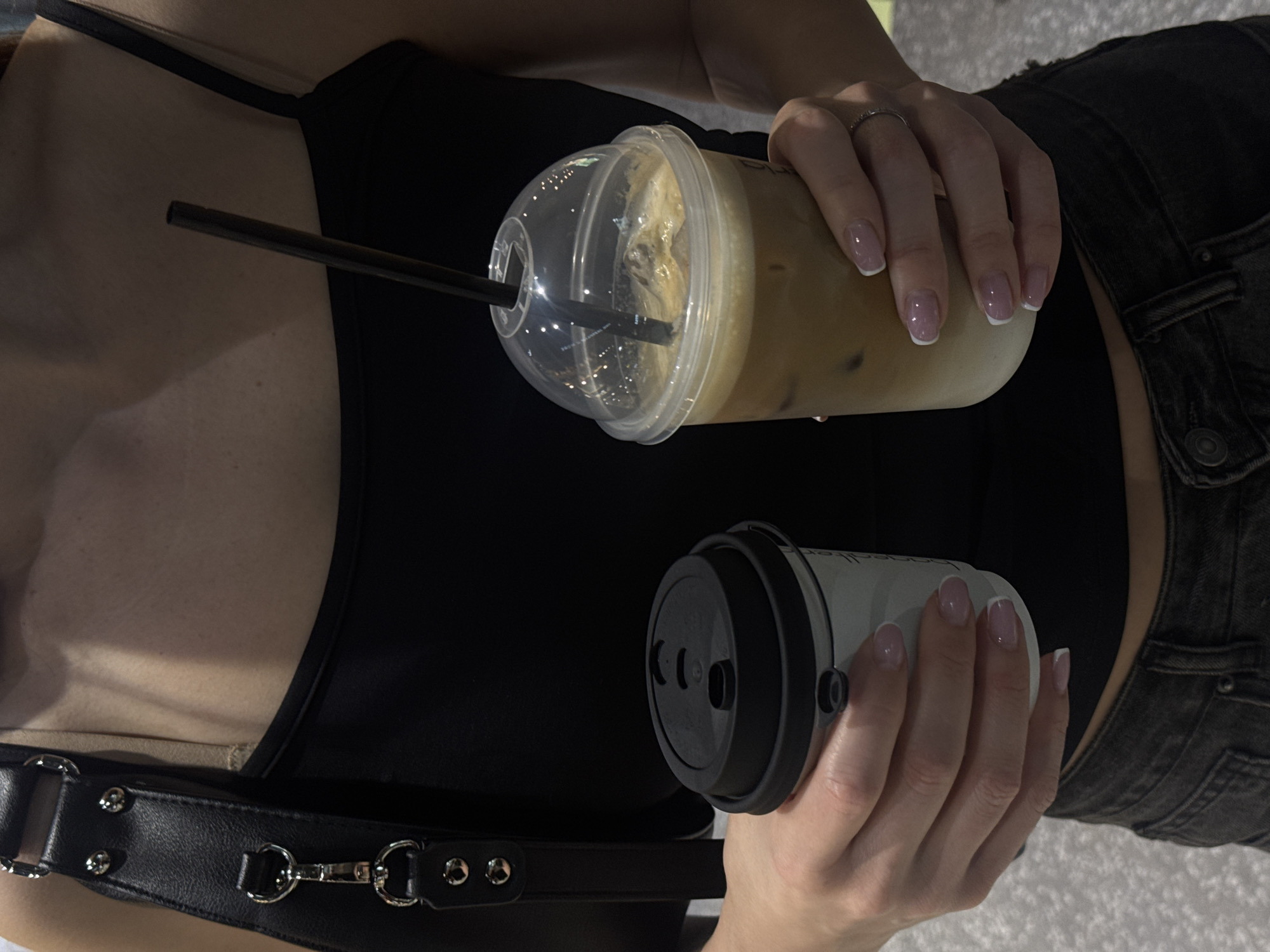 A woman is holding an iced coffee in a plastic cup with a lid and a black straw in her right hand and a coffee to-go cup with a black lid in her left hand. She is wearing a black top and dark-washed jeans. A black leather harness is visible over her chest. The location is unknown.