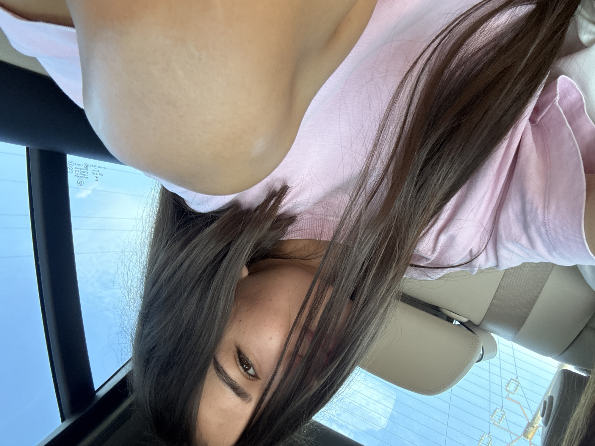 A young woman is in the back seat of a car, her dark hair falling across her face as she looks towards the camera. She is wearing a pink t-shirt. The interior of the car is visible, with beige seats and the headrest. Through the rear window, a bright blue sky with some clouds is seen, along with a glimpse of power lines. The image does not provide any information or context to determine a specific location within the city of Corbi, Romania.