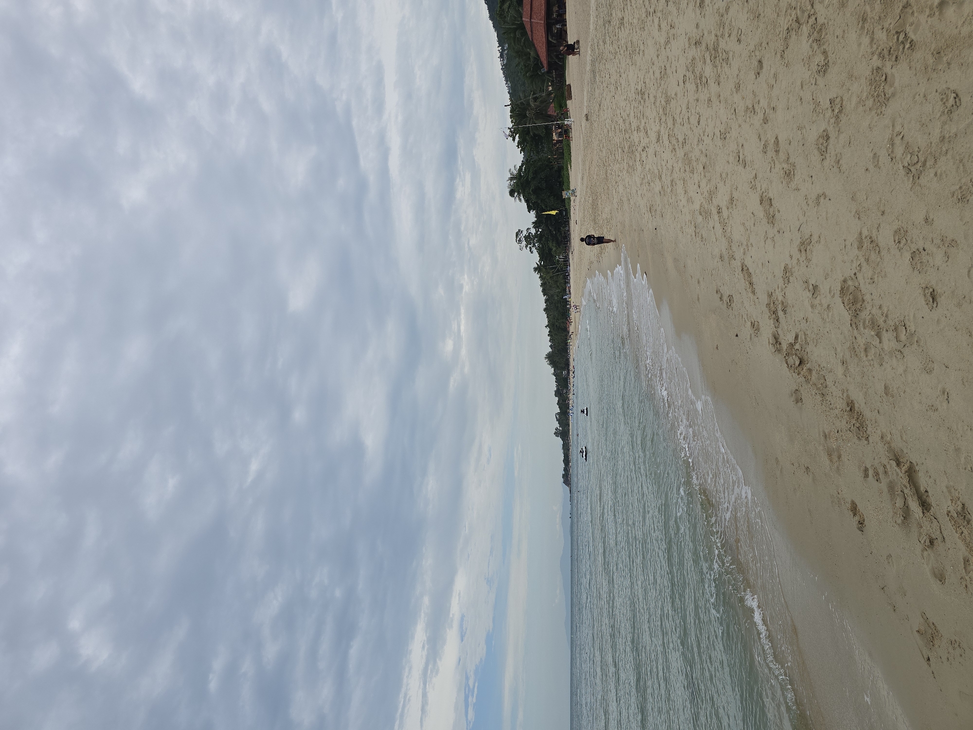 The image depicts a serene beach scene with a focus on a sandy coastline under a cloudy sky. The foreground is dominated by the textured sand, marked with footprints and a few small dunes. To the left, gentle waves lap at the shore, creating a white foam line. The water itself appears calm and a muted blue-green.
In the mid-ground, several people are visible. A lone figure, a person wearing a dark t-shirt and shorts, walks along the wet sand near the water's edge. Further out in the water, a few other individuals are scattered, some appearing to be swimming or engaged in water activities. Two jet skis are also present in the water, one moving and one stationary.
The background reveals a line of lush green palm trees and other vegetation, suggesting a tropical or subtropical location. A red-roofed structure, possibly a restaurant or resort, is nestled among the trees on the right side of the frame. In the far distance, faint outlines of hills or mountains can be seen against the horizon. The sky is filled with a layer of overcast, grey clouds, obscuring the sun and casting a diffused light over the scene. The overall impression is one of a peaceful, somewhat subdued beach day.
