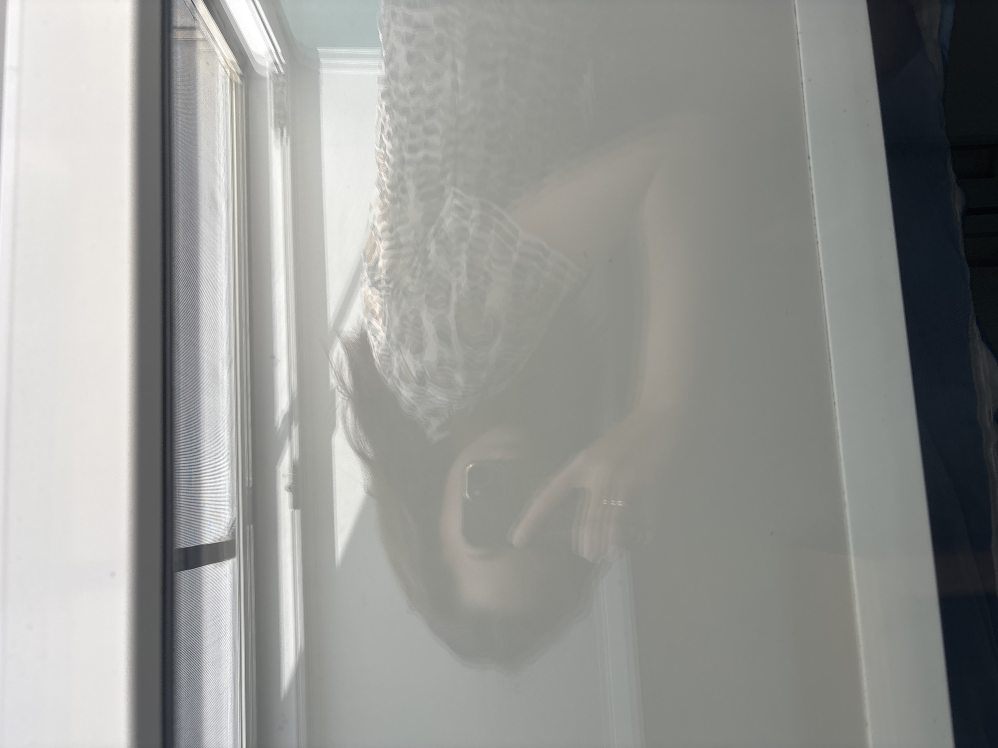 The image shows a translucent reflection of a woman holding a phone to take a selfie. She is positioned in the center of the frame, with her left hand raised to the phone, obscuring part of her face. She has long, dark hair and is wearing a patterned shirt. The reflection is cast against a diffused surface, possibly a frosted window or a sheer curtain, with light streaming in from the right side, creating shadows and highlights. The right edge of the frame shows a window with blinds. The scene appears to be indoors, and there is no information to suggest it is located in Inskoy, Russia.