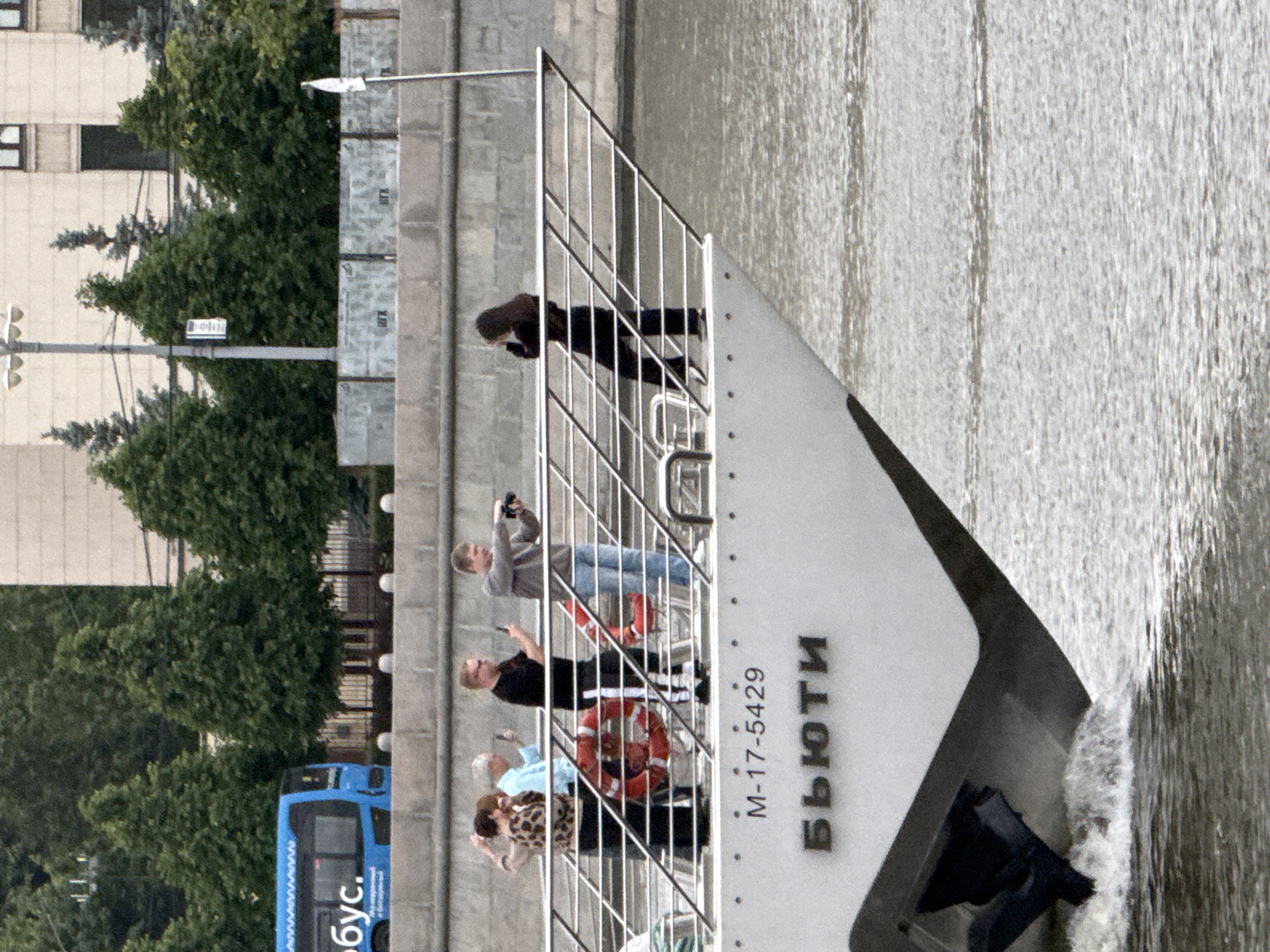 The image depicts a group of people standing on the deck of a boat. The boat is labeled with the name "Бьюти" (Beauty) and the registration number "M-17-5429." Some people are taking photos. The boat appears to be on a river, with a stone embankment and greenery visible in the background. A blue bus is also visible. This is likely the Moskva River in Moscow, Russia.