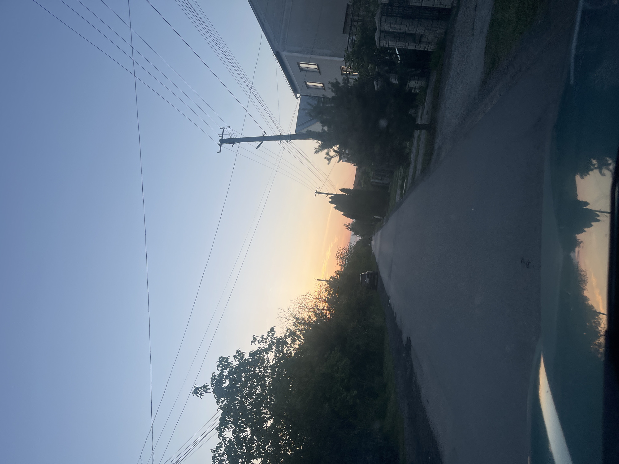 The image displays a tranquil rural or suburban road scene captured during either sunrise or sunset, indicated by the sky's gradient transition from light blue at the top to warm yellow and orange hues near the horizon. The photograph appears to be taken from within a vehicle, with a dark frame or window edge visible along the far right side.

A prominent wooden utility pole with numerous power lines stands near the center-left, with wires extending across the upper portion of the sky. On the left side of the paved road, a two-story building with several visible windows can be seen, partially obscured by trees and foliage. Various trees and bushes line both sides of the road, with many silhouetted against the bright horizon. The road itself, likely asphalt, extends into the distance, where a small car is parked on the shoulder, partially hidden by the surrounding greenery.

The scene depicts clear weather conditions with no clouds. The location is Storozhnytsia, Ukraine. No discernible activity or people are visible, and there is no text present in the image. The overall impression is one of peaceful observation of the changing light at the beginning or end of the day.