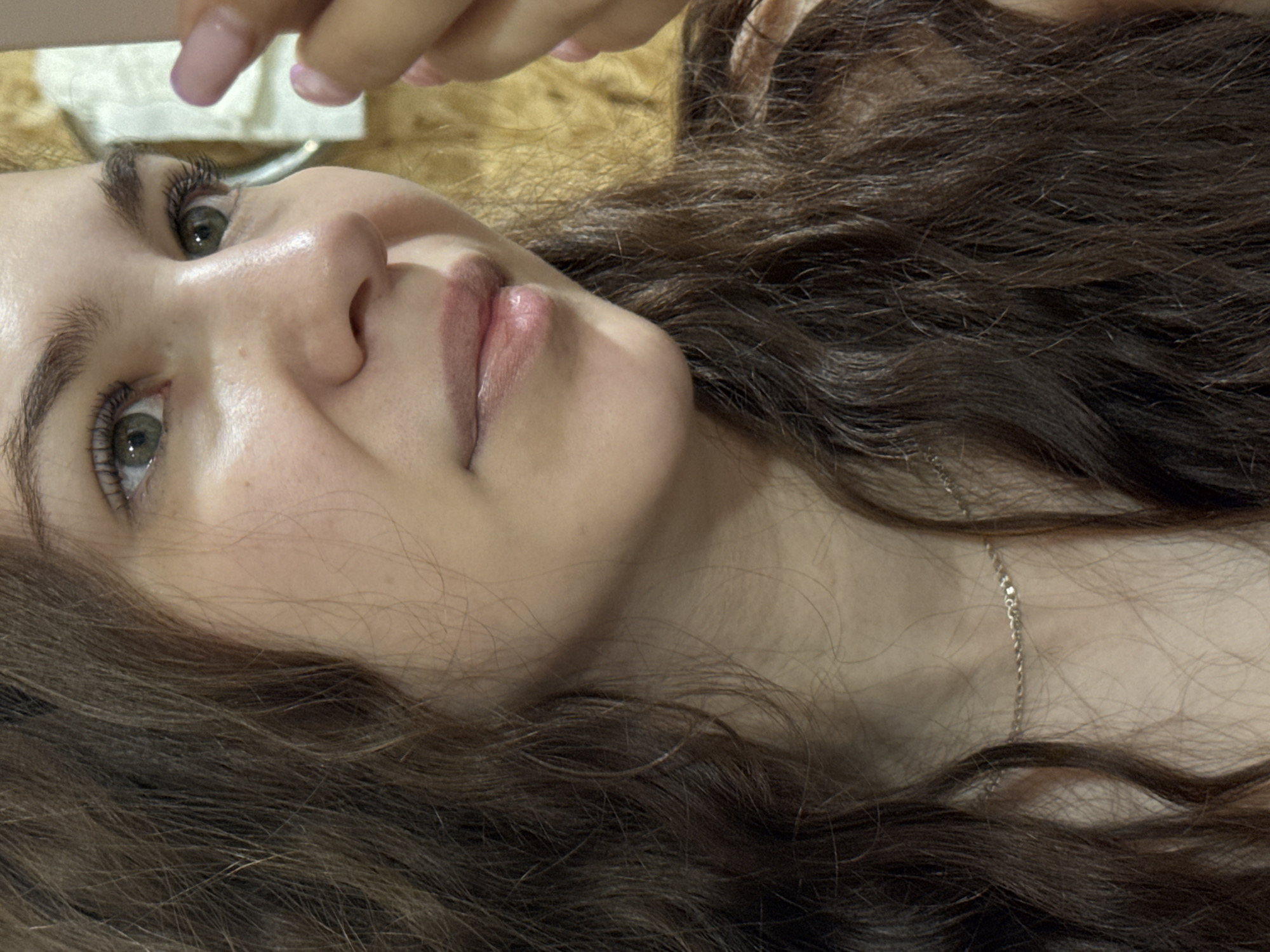A young woman with long brown hair and fair skin is lying down, looking up at the camera. She has light green eyes and a small silver necklace. Part of a hand with manicured nails is visible above her head. The background appears to be a wooden surface. There is no indication of Nizhnyaya Gar', Russia in the image.