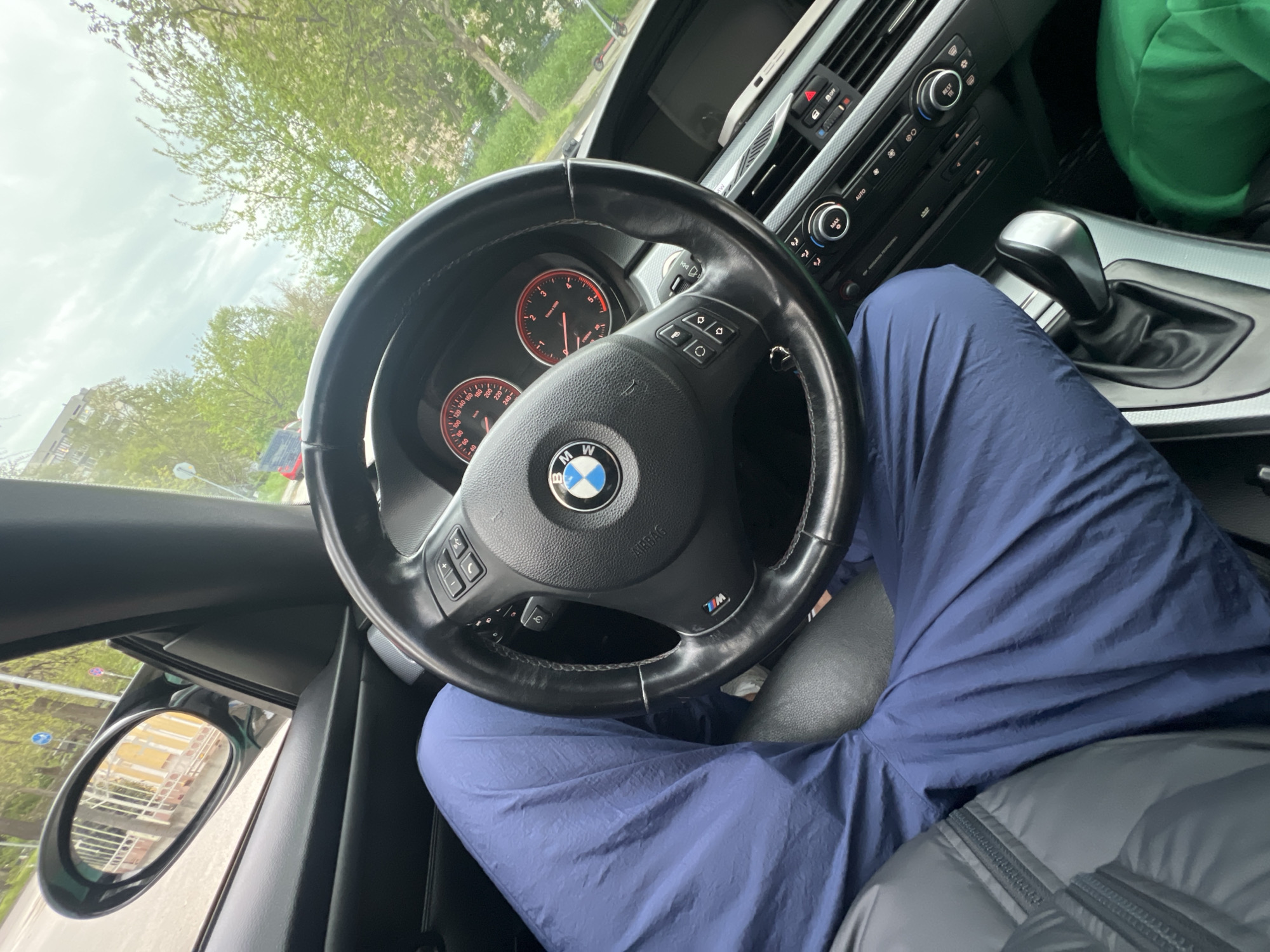 The image provides a driver's perspective from the interior of a BMW car, likely in Plovdiv, Bulgaria. The driver's legs, dressed in blue track pants, are visible beneath the prominent black leather steering wheel, which features the circular BMW logo at its center and "AIRBAG" text above it. A section of a dark grey puffy jacket rests on the driver's lap.
Through the windshield, an overcast sky and vibrant green trees with fresh leaves are visible, indicating daytime. The side mirror reflects a light-colored building with multiple windows. The dashboard is detailed, showing an instrument cluster with red-lit gauges displaying numerical values such as "60," "40," "180," "200," "220," and "240" for speed, and "2," "3," "5" for RPMs. The center console includes climate controls with buttons labeled "AUTO" and "REST," along with a distinct automatic gear shifter. Additional text on the steering wheel includes "MW" near the bottom right. The overall scene suggests the car is either parked or stationary.