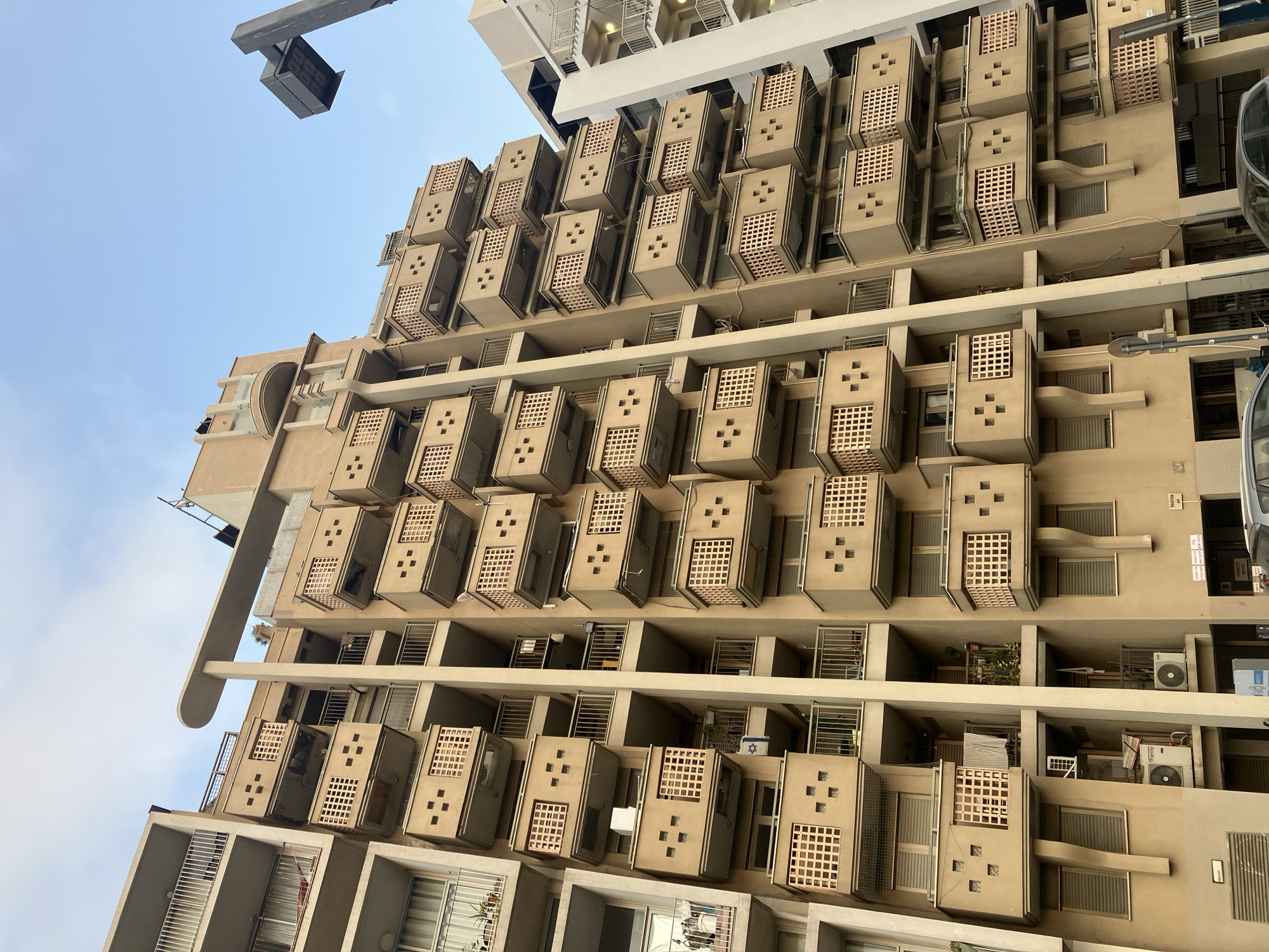 An exterior, eye-level, low-angle shot features the facade of a contemporary apartment building with a beige-colored exterior. The building has numerous balconies, each with a distinctive lattice-work design and small, square cutouts. The balconies protrude from the building at various levels and are evenly spaced. A thin, white vertical beam runs up the center of the building's facade. The sky above is a clear, pale blue. To the right, a dark grey, modern streetlamp is visible, positioned against the sky. The bottom of the frame shows the street level, including a dark grey car, a black car, and portions of store entrances with signs in Hebrew. The architectural style is modern, with clean lines and geometric shapes. The building appears to be in an urban environment, likely Tel Aviv, Israel, given the Hebrew signage and architectural style. No people are visible in the frame.