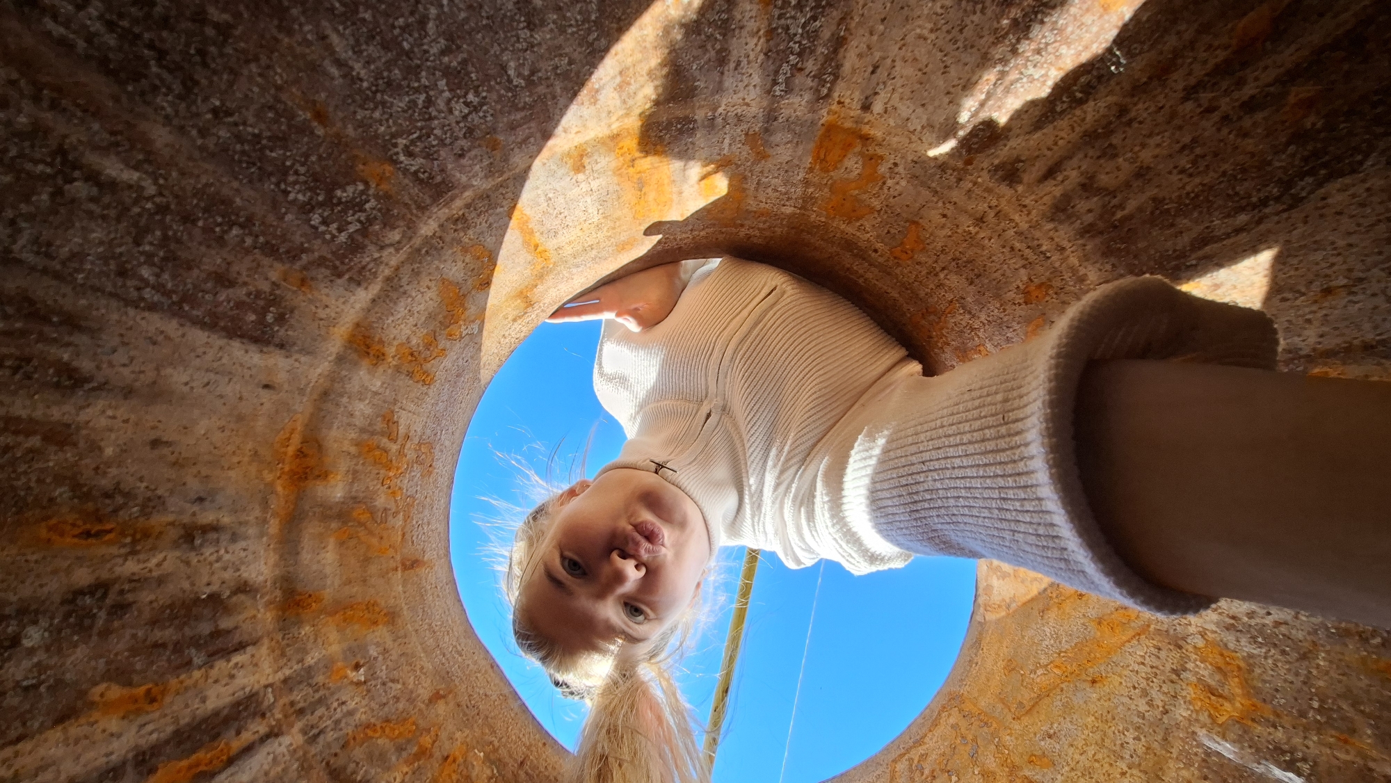 A wide-angle, low-angle shot captures a young woman peering down into a large, rusty pipe or cylinder. Her face, centered within the circular opening, displays a playful "fish lips" or "duck face" expression. She has blonde hair, pulled back into a ponytail, and is wearing a light-colored (likely white or cream) ribbed turtleneck sweater.
The bright blue sky serves as the background, visible through the opening and framing her head and shoulders, suggesting a clear, sunny day. Sunlight casts dynamic highlights and shadows on the rough, textured interior walls of the pipe, which are heavily encrusted with rust, displaying various shades of brown, orange, and grey. The perspective implies the camera is positioned inside the pipe, looking upwards at the woman. This image captures a lighthearted moment of interaction within a weathered industrial or natural structure, located in Большой Букор, Russia. No visible text is present.