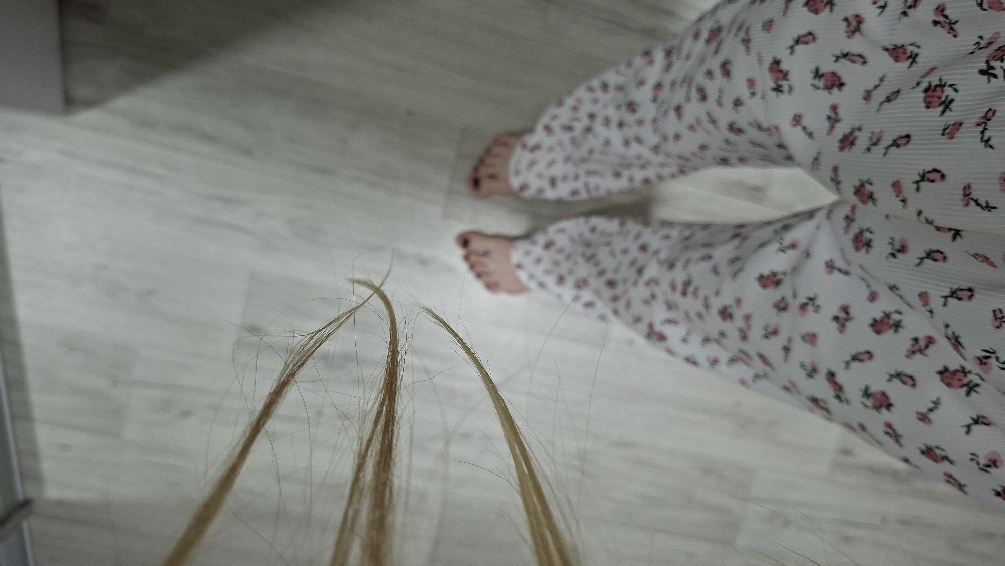 The image depicts a first-person, downward-facing view of a person's lower body and the immediate environment in the city of Chaikovsky, Russia. The individual is wearing white pajama-style pants patterned with small pink roses and green stems. Below the pants, bare feet are visible, with all visible toenails painted black. To the left side of the frame, multiple strands of long, light-colored hair, appearing blonde or light brown, are hanging or resting, with some individual hairs scattered on the light-colored wooden or laminate floor. The floor consists of distinct planks. A blurred light-colored vertical surface, possibly a wall or furniture, is visible in the upper background.