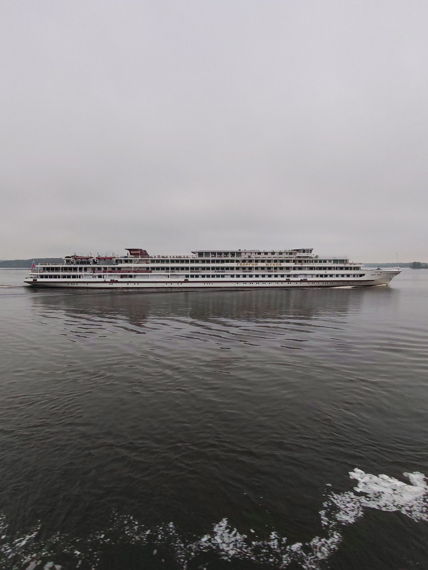The image captures a large, white river cruise ship sailing on a body of water under an overcast sky, potentially located in Kalyazin, Russia. The ship has multiple decks with numerous windows and possibly balconies, and what appears to be a Russian flag is visible on the stern. The water is choppy, with visible waves and white foam near the bottom right corner, likely indicating the wake from a moving vessel. In the background, a strip of land can be seen on the horizon. The overall scene appears calm but muted due to the weather conditions.