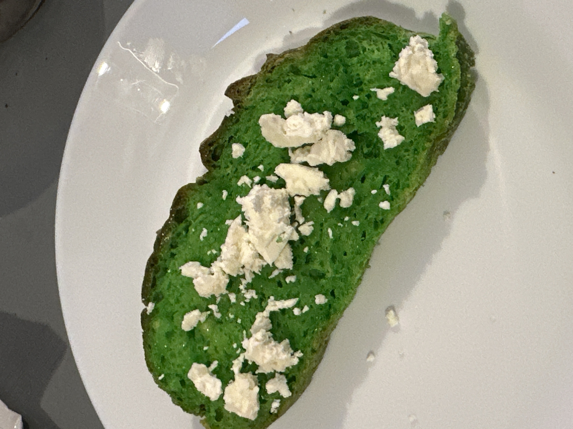 A single slice of green-colored bread, topped with crumbled white cheese, rests on a white plate. The bread appears to be a dense, possibly sourdough, loaf, with its vibrant green hue likely achieved through food coloring or ingredients like spinach. The cheese is scattered unevenly across the surface of the bread, with some pieces larger and more prominent than others. The plate itself is simple and white, with a slight sheen reflecting light from above. The background is blurred, suggesting a shallow depth of field, but hints at a dark, textured surface, possibly a table. There is no indication of the specific location in Moortown, Ireland, nor any people or actions occurring in the scene.