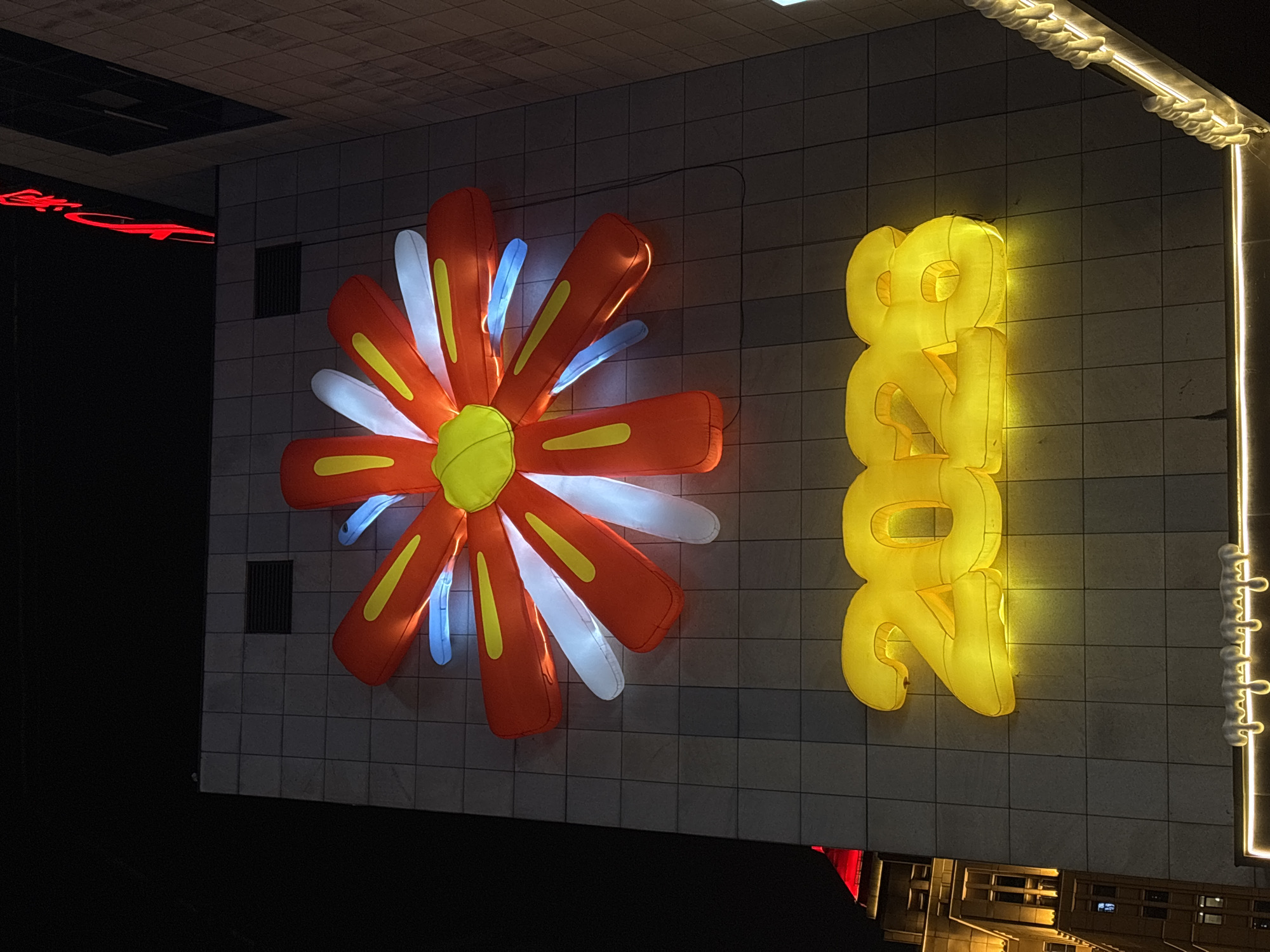 The image depicts an outdoor setting in Yuzhong District, China, featuring illuminated decorations on a tiled wall. To the left is an oversized, stylized flower decoration with orange petals lined with yellow, interspersed with white and blue accents. To the right, vertically arranged yellow numerals, "2024", likely made of inflatable or soft material, are illuminated against the wall. An elongated white lighting fixture with a decorative pattern runs along the wall's right edge. Below, the faint reflection of buildings is visible. The setting appears to be at night, adding emphasis to the bright, glowing display.