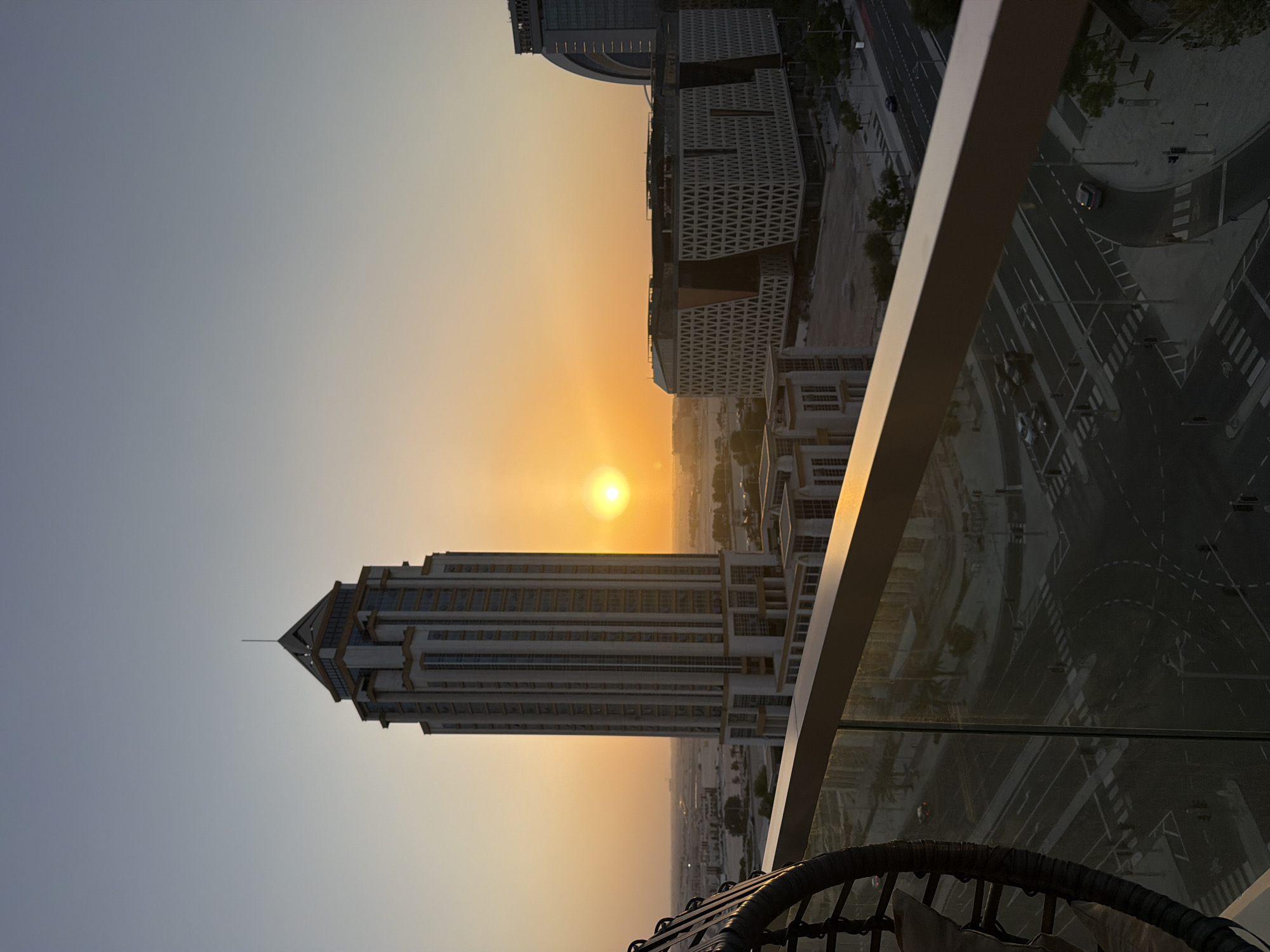 The image is a vertical, eye-level shot of a city skyline at sunset. The sun is a bright orb in the center-left of the frame, casting a warm, orange glow across the sky. A tall, modern skyscraper dominates the foreground, its glass facade reflecting the sunset. To its right, other buildings and a street scene with cars are visible. A glass railing and the edge of a woven chair are in the bottom right corner, suggesting the vantage point is from a balcony or a similar elevated location. The sky is a gradient of pale blue at the top, transitioning to a soft orange near the horizon. The overall mood is serene and contemplative, with the setting sun creating a peaceful atmosphere. There is no visible text in the image.
