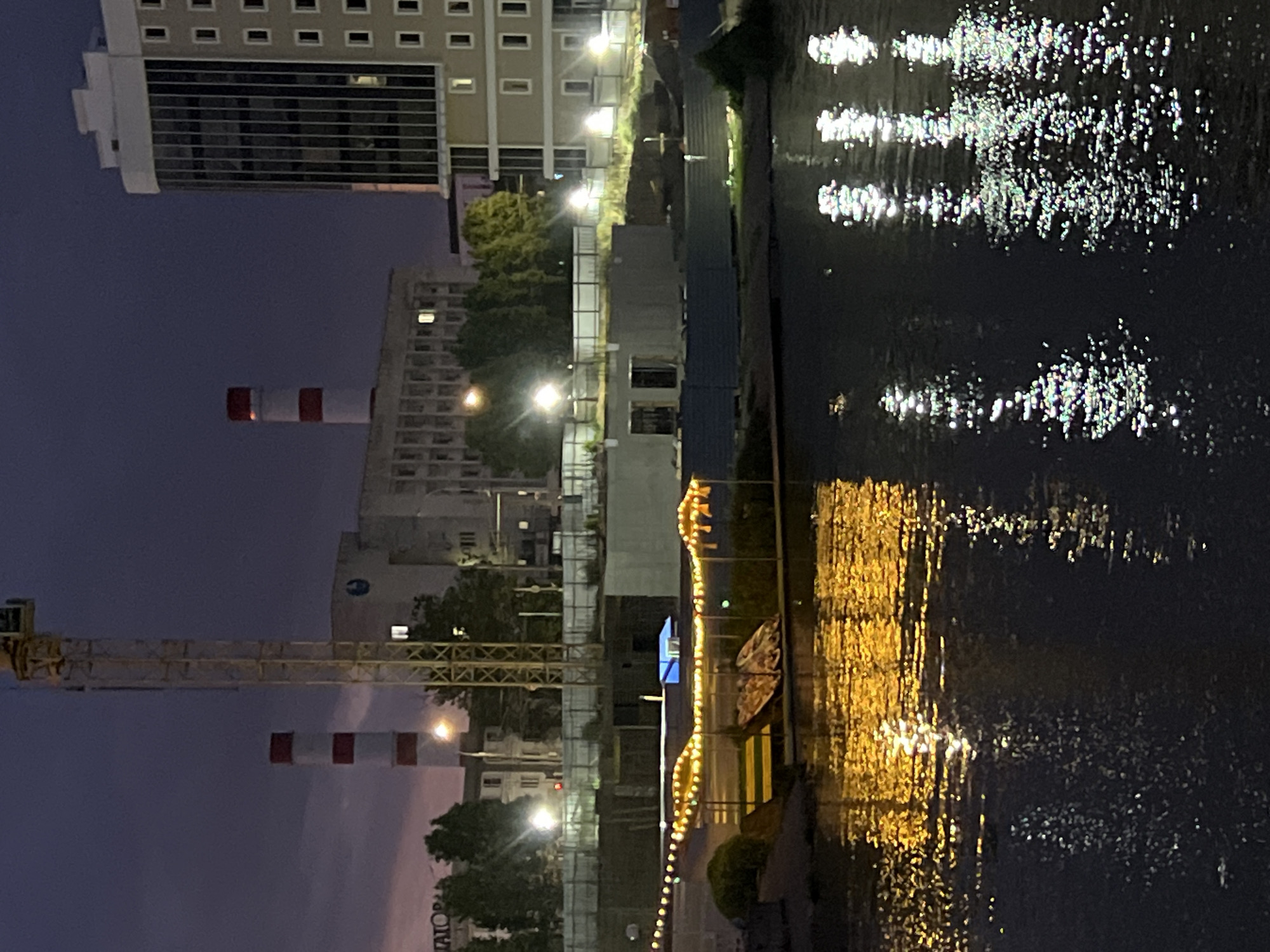 This is a night-time outdoor shot of a riverfront area in Krasnodar, Russia. The image captures a scene with industrial elements, a modern building, and a body of water reflecting lights.
In the background, a tall yellow construction crane is prominently visible. To the left of the crane, two smokestacks with red and white bands rise against the dark sky. Behind them, a grey industrial building complex with multiple windows is seen. To the right, a modern, multi-story building with a glass facade and numerous square windows dominates the skyline. The building appears to be lit from within, with some windows glowing.
In the foreground, a body of dark water stretches across the bottom of the frame. The water reflects the lights from the scene, creating shimmering patterns of yellow and white. A strip of land is visible along the edge of the water, with a structure decorated with string lights. Two boats are also present on this land. The area appears to be under construction, with fencing and building materials visible in the middle ground. Several bright streetlights illuminate parts of the construction site and the surrounding trees. The overall impression is one of urban development and industrial activity by a waterfront at dusk or night.