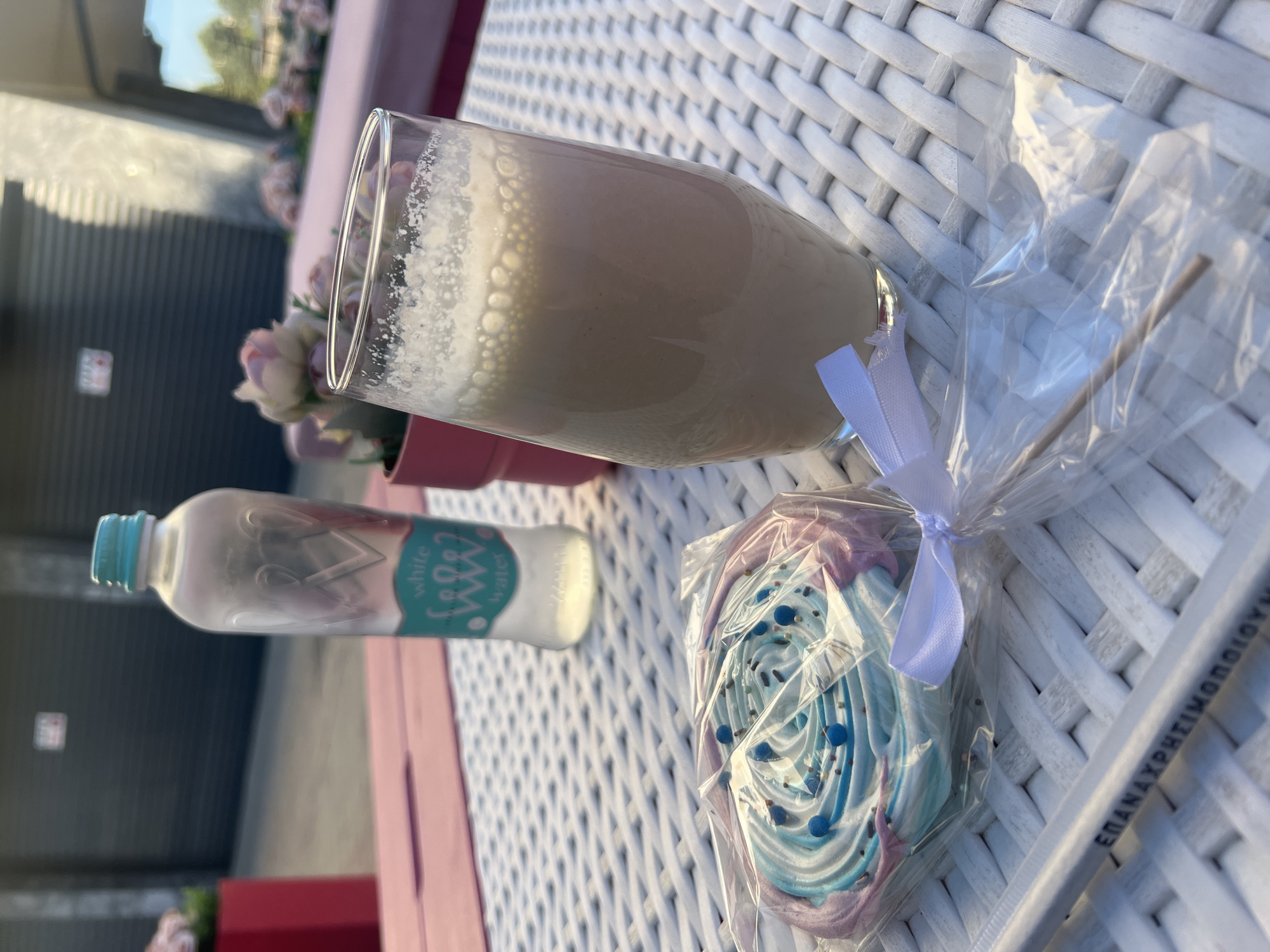 A close-up shot taken outdoors, likely on a cafe or restaurant patio, shows a set of items on a white, woven-style table. A tall glass filled with a light-colored drink, likely a latte or milkshake, sits prominently. Next to it is a small bottle of "white water" from Devina, indicating the scene is in Bulgaria. Beside the glass, there's a wrapped lollipop featuring pastel colors and sprinkles, tied with a white ribbon. In the lower right, part of a white paper straw with Greek lettering is visible. The background shows hints of flowers and other patio furniture, suggesting the setting is a cafe or similar establishment, potentially in Plovdiv, Bulgaria given the water bottle brand. There are no people visible in the frame, focusing solely on the food and drink items. The angle of the photo is slightly sideways, implying the shot was taken from a position at the table.