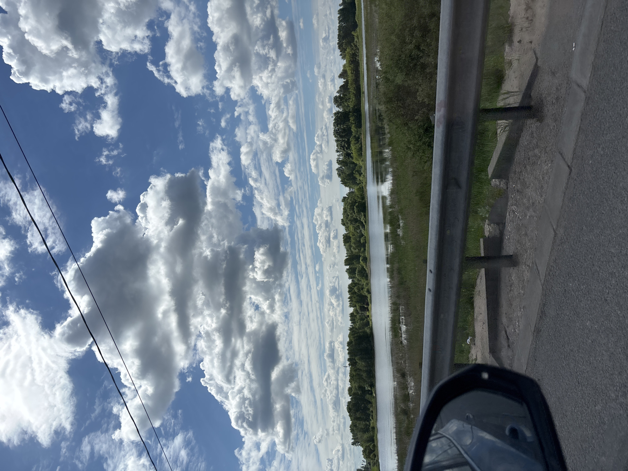 This image captures a vibrant outdoor scene from the perspective of a vehicle, likely in motion. The dominant feature is the expansive sky, showcasing numerous large, fluffy white cumulus clouds against a bright blue background. Some darker, denser clouds are also present, adding depth to the sky. Two dark power lines diagonally cross the upper left part of the frame.
Below the sky, a landscape featuring a road, guardrail, and a body of water unfolds. On the right side, an asphalt road is visible, running alongside a sturdy grey metal guardrail supported by concrete posts. Beyond the guardrail, green grass and other vegetation line the edge of a calm body of water, possibly a river or canal, which stretches horizontally across the midground. The water beautifully reflects the sky and clouds, creating a striking mirror effect. A dense line of dark green trees flanks the far bank of the water body, extending into the distance.
In the bottom right corner, a portion of a car's side-view mirror is visible, reflecting elements of the vehicle and its surroundings. A small, white bird is perched on the ground near the guardrail, situated between it and the water.
The scene depicts bright daylight, indicative of a clear and sunny day with scattered clouds. The overall impression is one of peaceful transit through a scenic, likely rural or semi-rural, area, consistent with the location context of Staroye Melkovo, Russia. No visible text is present in the image.