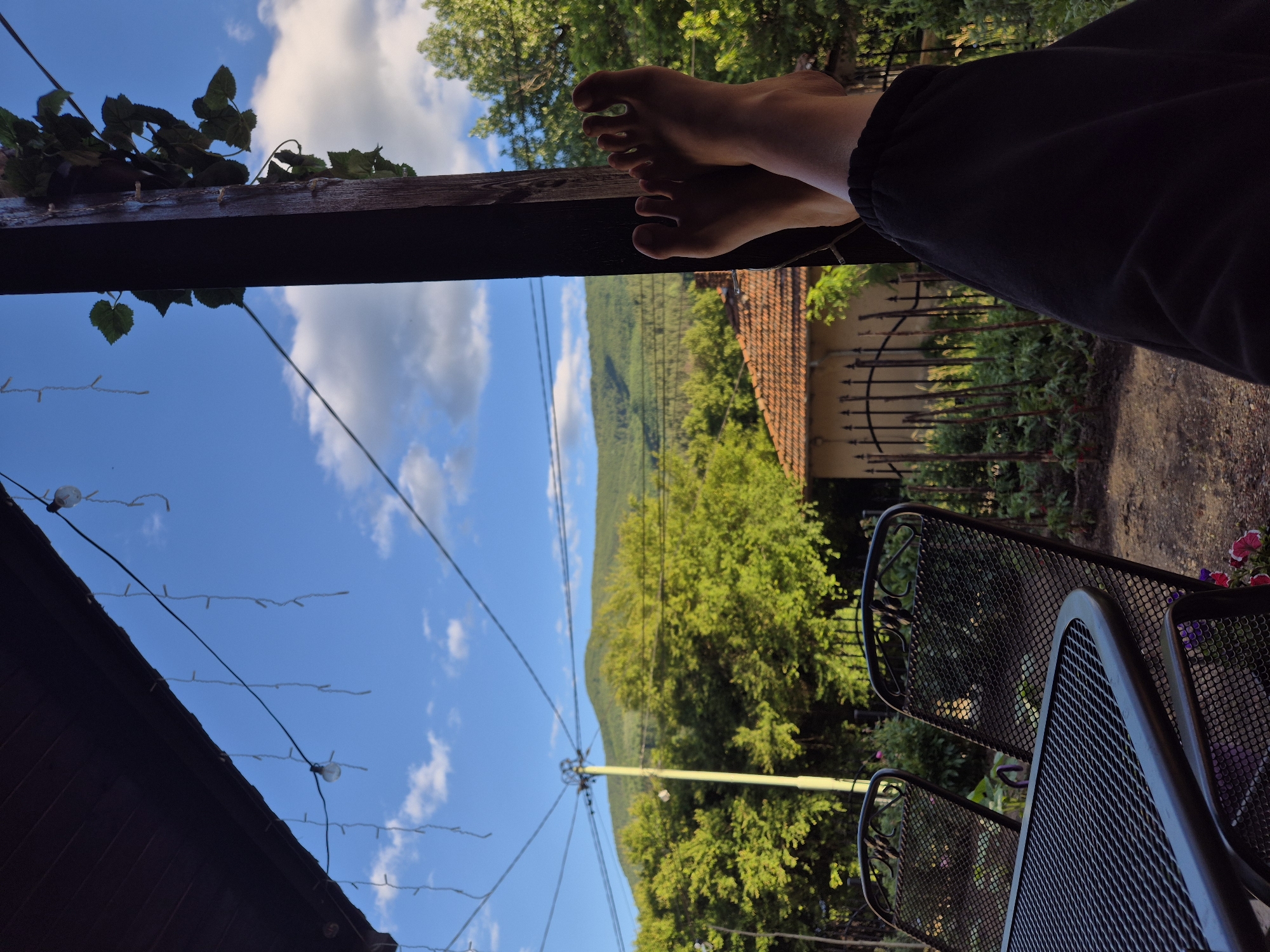 The image depicts a perspective from a covered outdoor seating area, likely a porch or patio, in Iskrets, Bulgaria. In the foreground, a person's bare feet are propped up on a dark-colored wooden post, wearing black sweatpants. The feet are illuminated by natural light, highlighting the skin tone and toenails. Two dark, metal mesh chairs and a matching table are visible to the left, suggesting a dining or relaxation setup.
The background reveals a rural landscape. Lush green trees dominate the mid-ground, with a traditional house featuring a terracotta tiled roof and a rustic fence visible behind the chairs. Beyond the trees, a gentle, green mountain slopes upward against a bright blue sky scattered with white clouds. String lights, some illuminated and some not, hang from the covered area, adding a decorative element. The overall impression is one of a peaceful, outdoor setting, possibly during the late afternoon or early evening, given the quality of light and shadow.