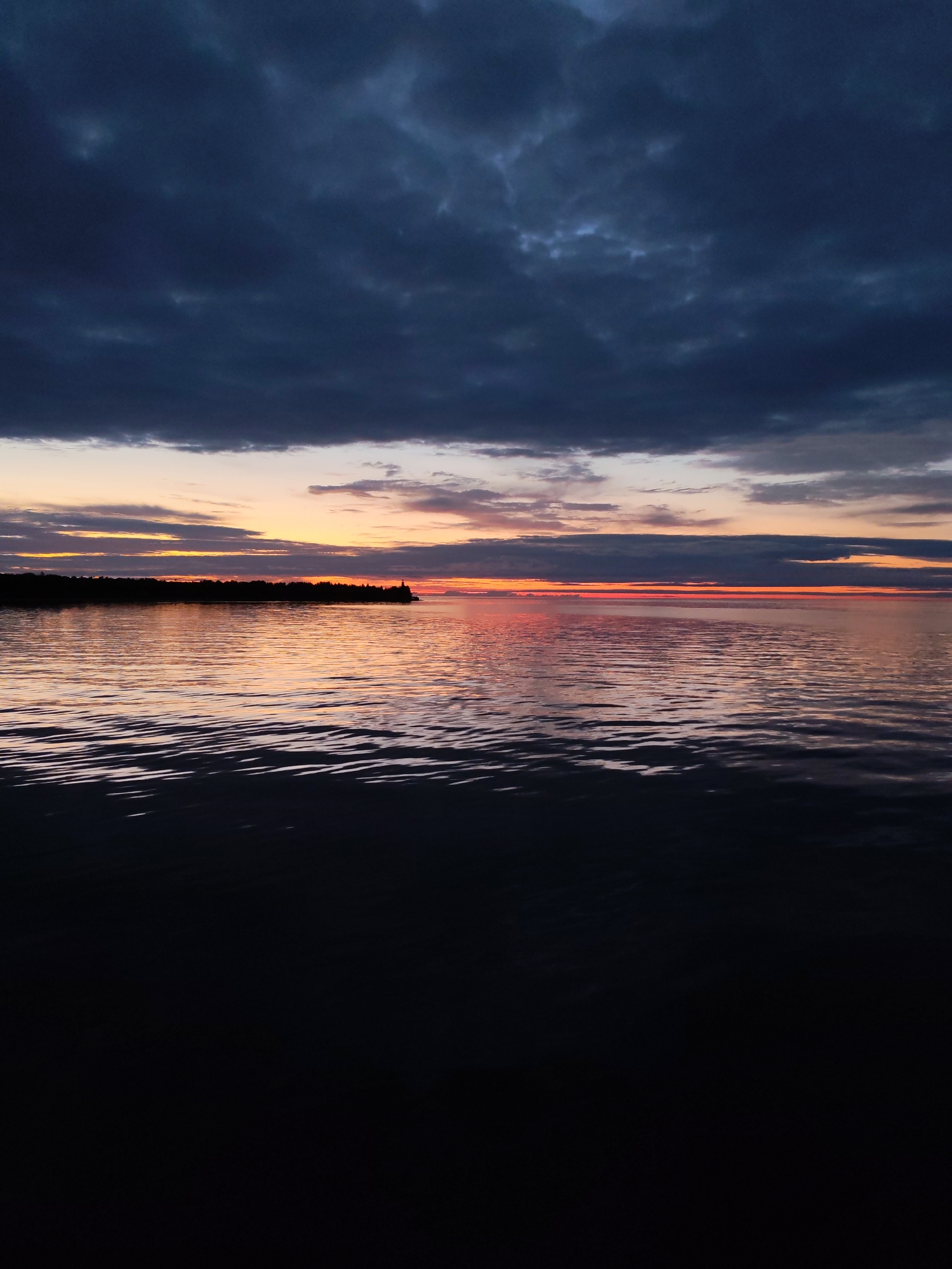 This is a photograph of a body of water at sunset. The sky is filled with dark, dramatic clouds in the upper portion of the frame, transitioning to bands of orange, pink, and yellow near the horizon. The water reflects the colors of the sky, creating a shimmering surface. In the distance, a dark silhouette of trees lines the horizon, with a small structure resembling a lighthouse visible on a point of land. The foreground of the water is darker and appears to be a deeper, calmer expanse. The image captures a serene, natural landscape scene, likely taken from a boat or the shore. The location is Shcheleiki, Russia.