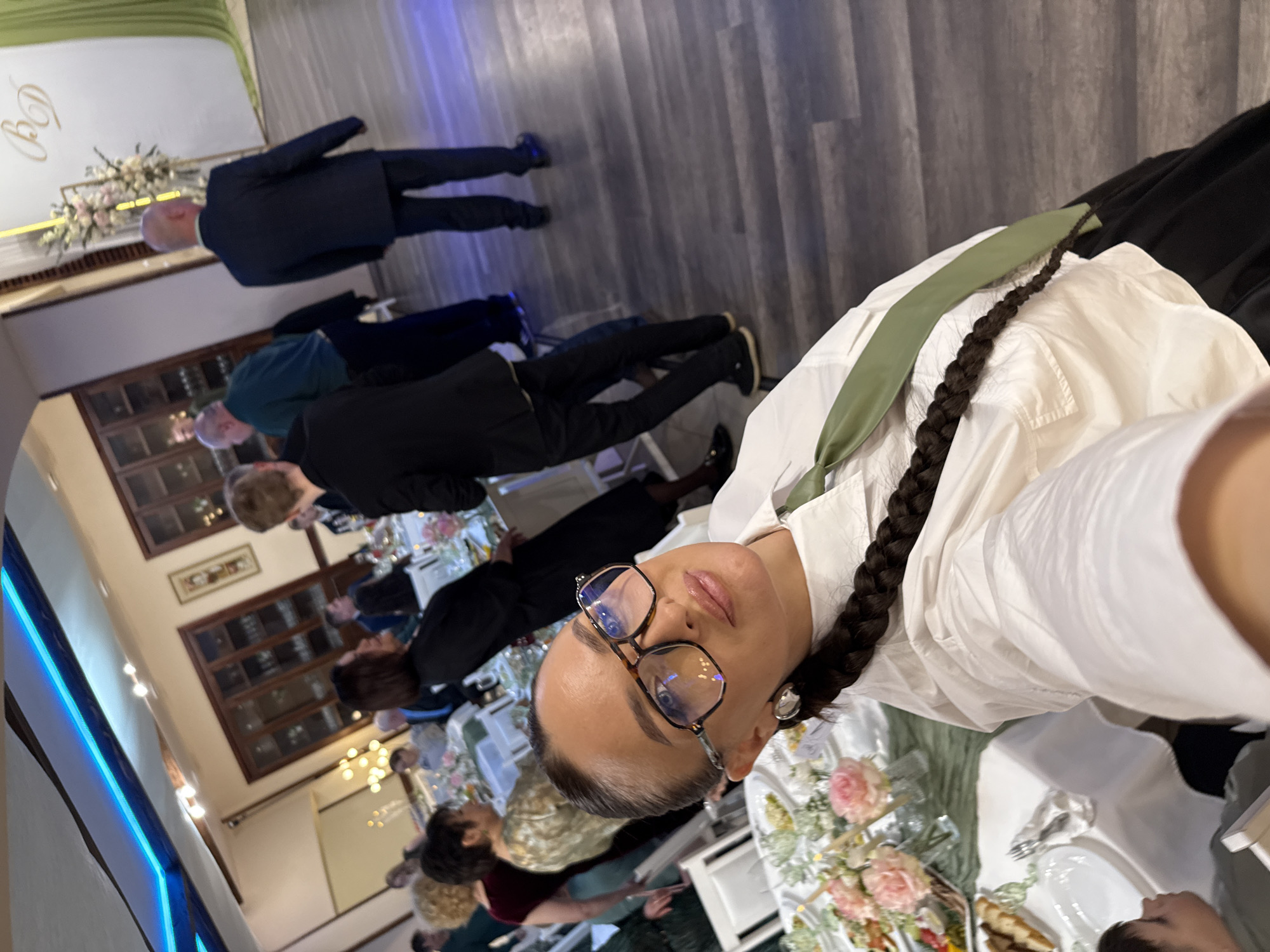 The image appears to be a selfie taken at a wedding reception. The person taking the selfie is wearing glasses, a white shirt, and a green tie, with their hair in a braid. The background shows a long table set for a meal, with flowers, dishes, and cutlery. People are seated at the table, including individuals in suits and dresses. The reception is taking place indoors, possibly in a banquet hall or event space, as suggested by the formal table setting and lighting. The presence of Russian text (likely "BD") on a banner suggests the event might be in Russia, and according to the information in the prompt, it may take place in the city of Chaikovsky.