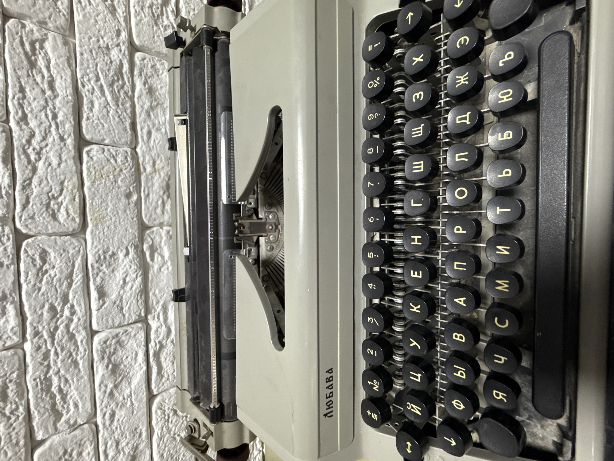 The image shows a close-up, angled view of a vintage typewriter with Cyrillic characters on the keys. The typewriter is light gray in color with black keys. The name "ЛЮБАВА" (Lyubava) is printed vertically on the machine's body. The background is a white brick wall. It is not possible to determine the location of the image being in Votkinsk, Russia or not.