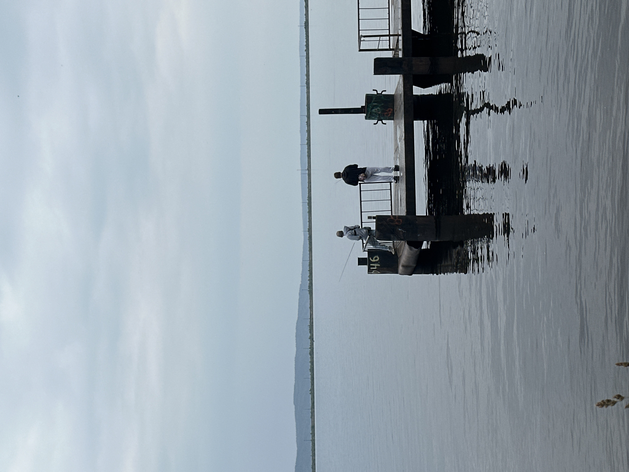 Two individuals are fishing from a pier on a body of water under an overcast sky. The pier is made of concrete and metal, with railings on one side. The water is calm, reflecting the sky and the pier. In the background, a distant treeline is visible on the horizon. One person is standing closer to the viewer, wearing a dark hooded sweatshirt and light-colored pants, holding a fishing rod. The other person is further away, wearing a light-colored hooded jacket and dark pants, also fishing. The pier has some graffiti on it. The overall scene is muted and serene, suggesting a cool or cloudy day.